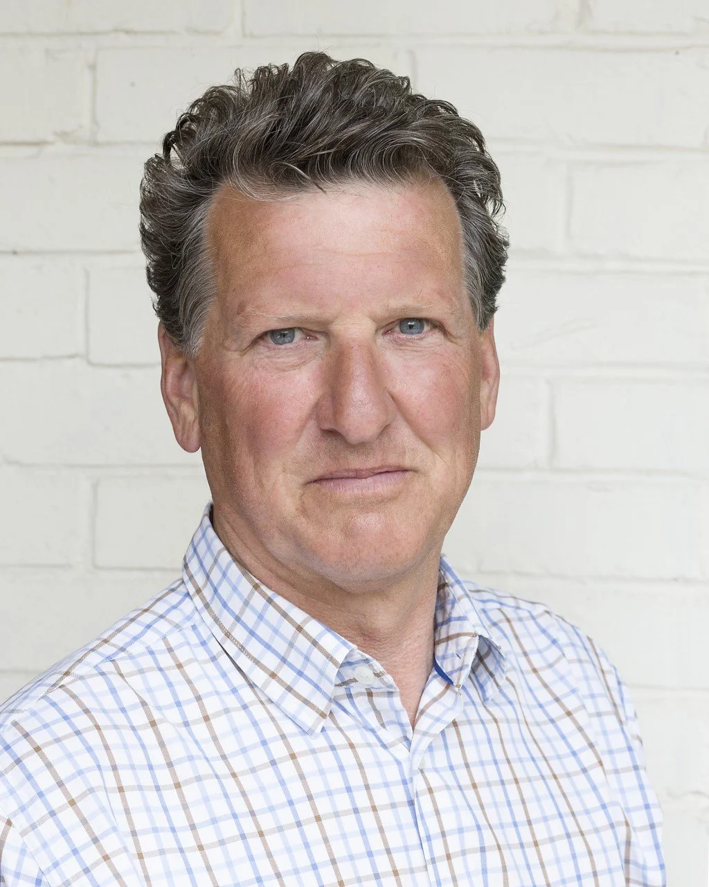 A middle-aged man with light skin, wavy short dark hair, and blue eyes, wearing a checkered shirt, standing in front of a white brick wall.