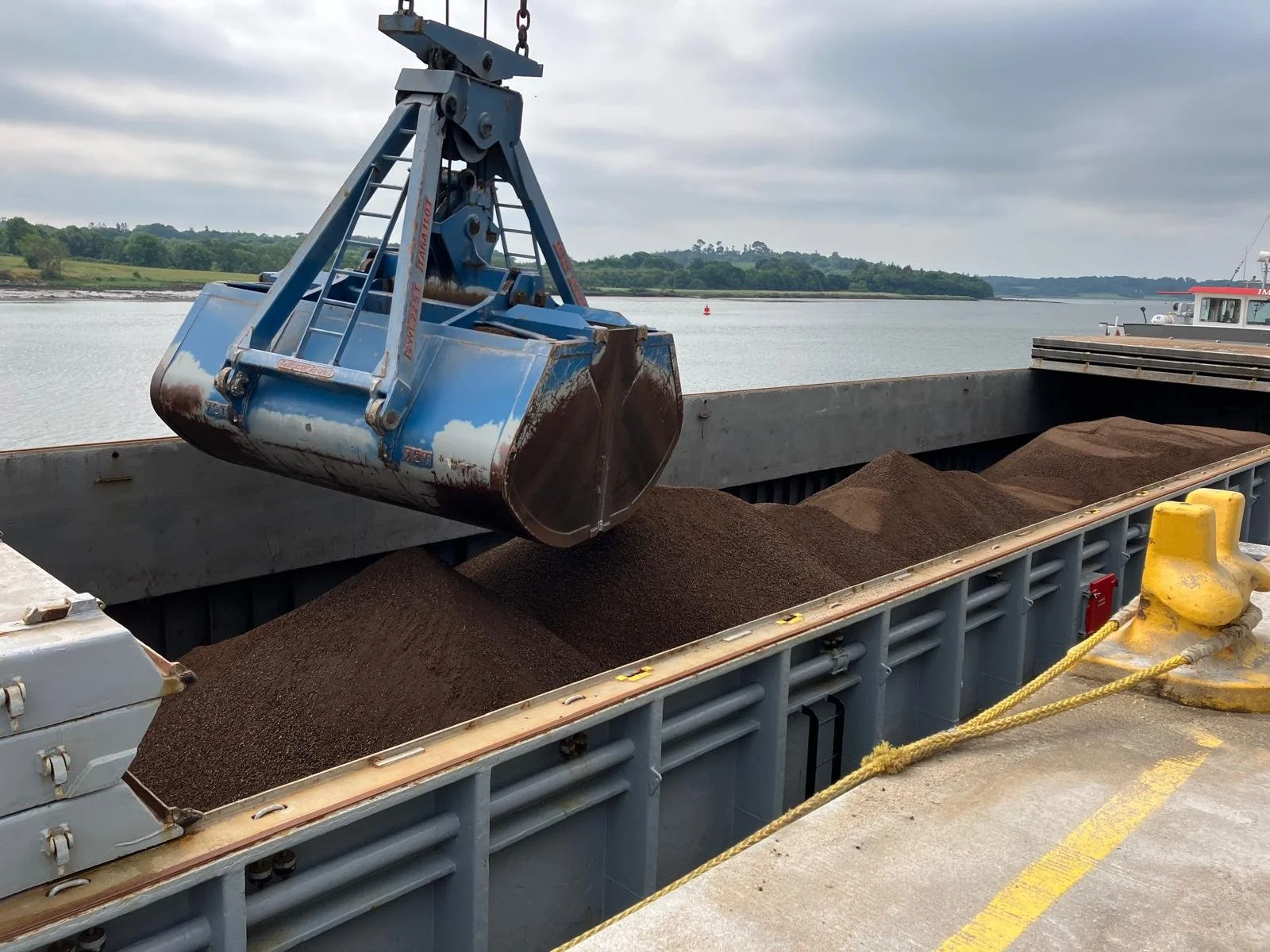 A cargo ship's open container filled with a commodity, with a large mechanical grab bucket suspended above it by a crane, on a body of water with a distant shoreline and cloudy sky in the background.