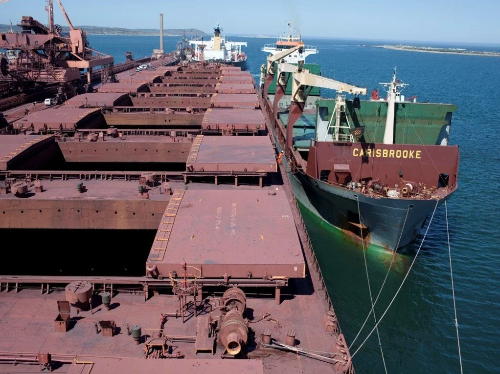 View of a large cargo ship moored at a dock, with cargo holds and cranes visible on deck, and a green and red hull with the name "Carisbrooke" on the bow, on a calm sea with land in the background.