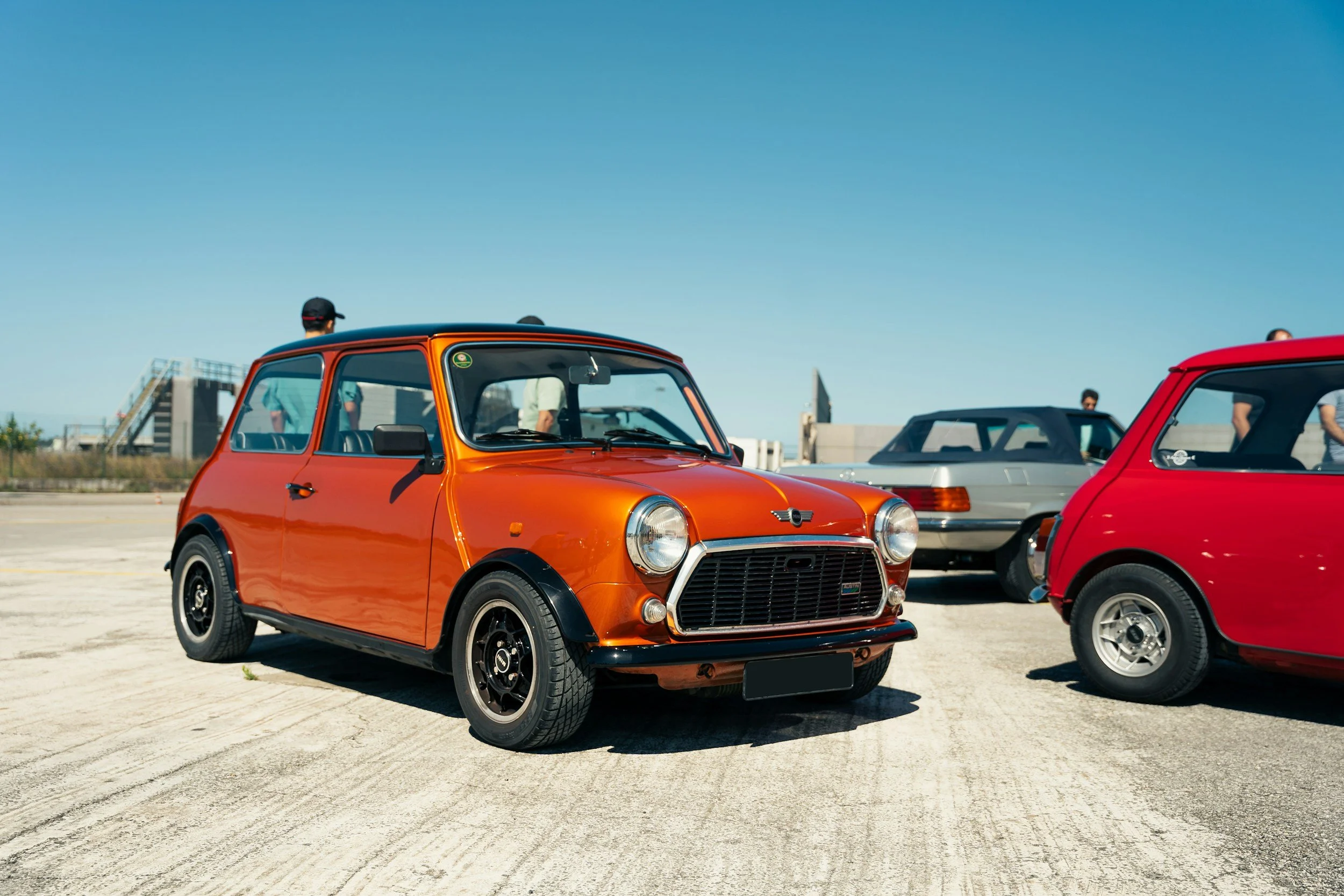 A vintage orange Mini Cooper parked on a rooftop parking lot with other classic cars and people in the background.