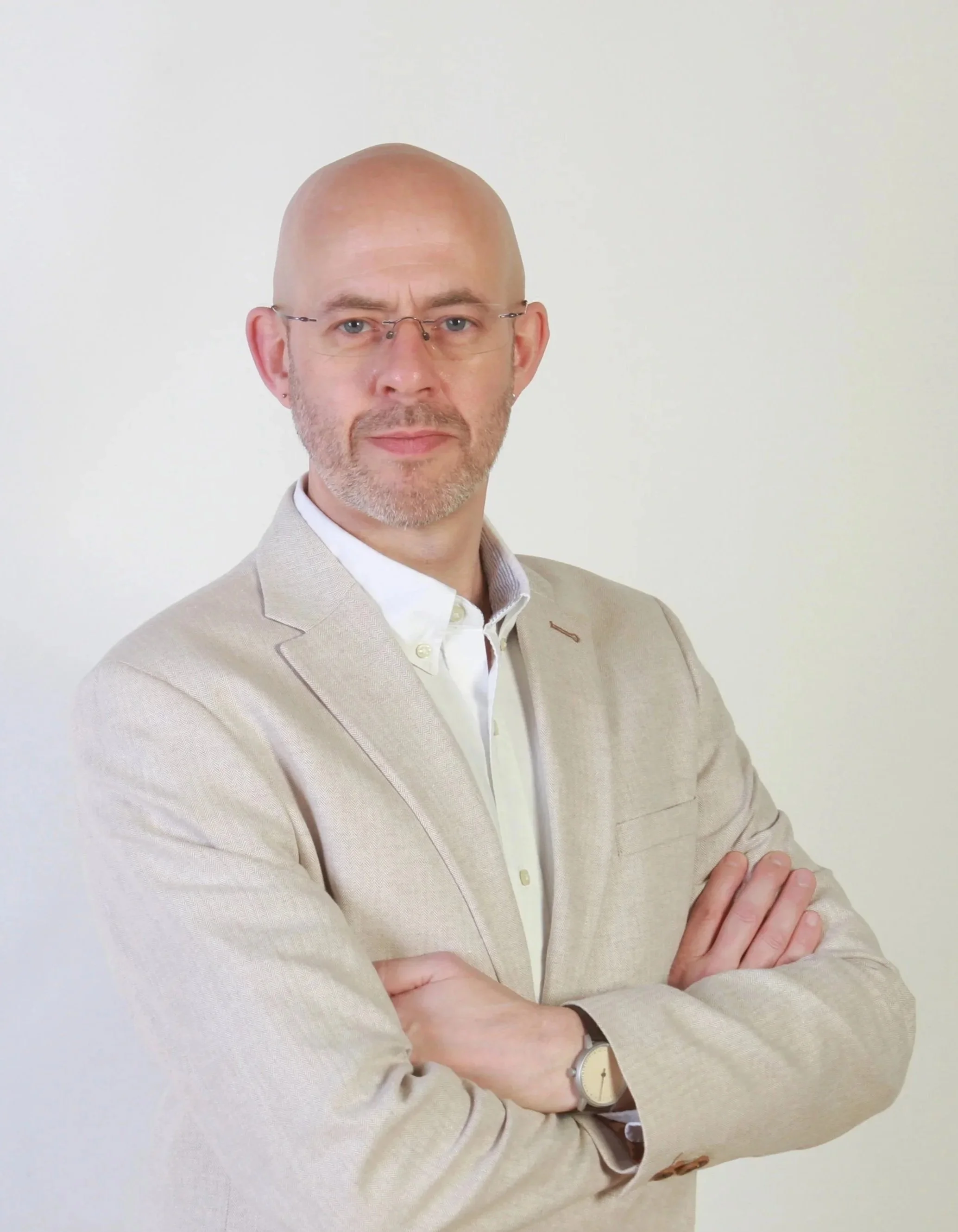 A picture showing Michael Wood, a middle-aged man with glasses and a beard, wearing a beige blazer and a white shirt, standing with arms crossed against a plain white background.