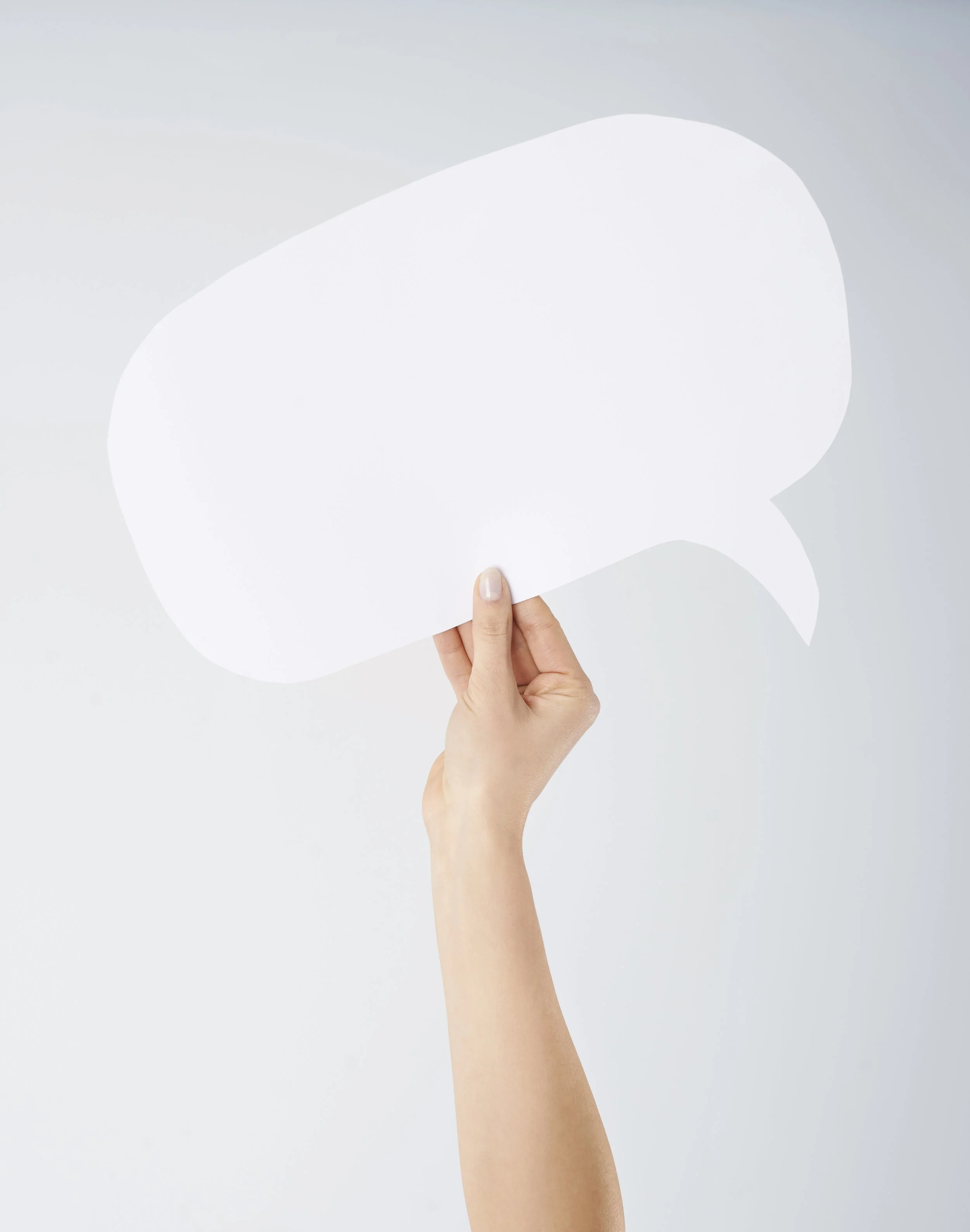 A hand holding a blank white speech bubble cutout against a light gray background.