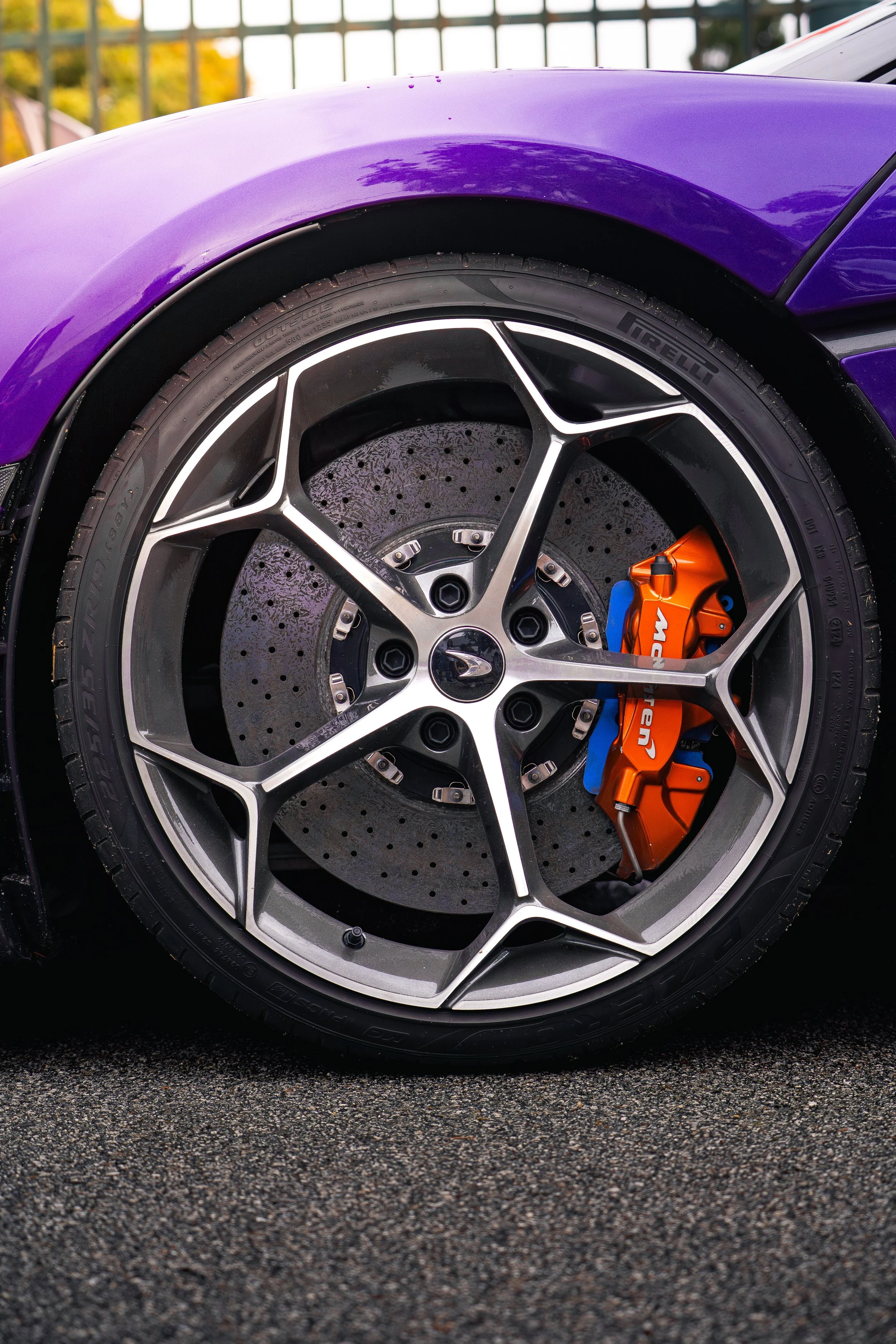 Close-up of a sports car tire, rim, and brake caliper. The rim has a modern design with silver and black accents. The brake caliper is orange with blue accents and branding.