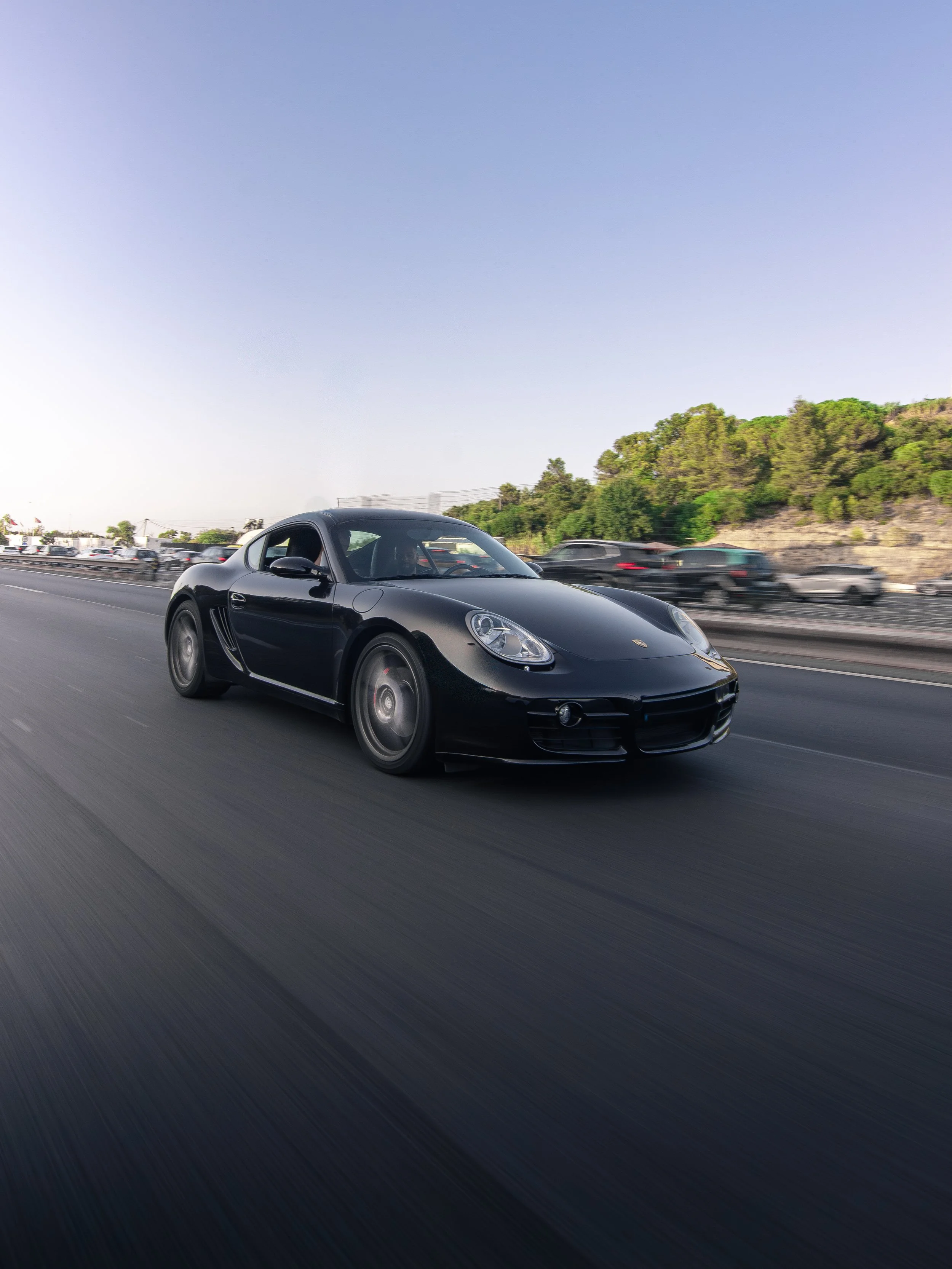 Black Porsche sports car driving on a freeway with cars and greenery in the background.