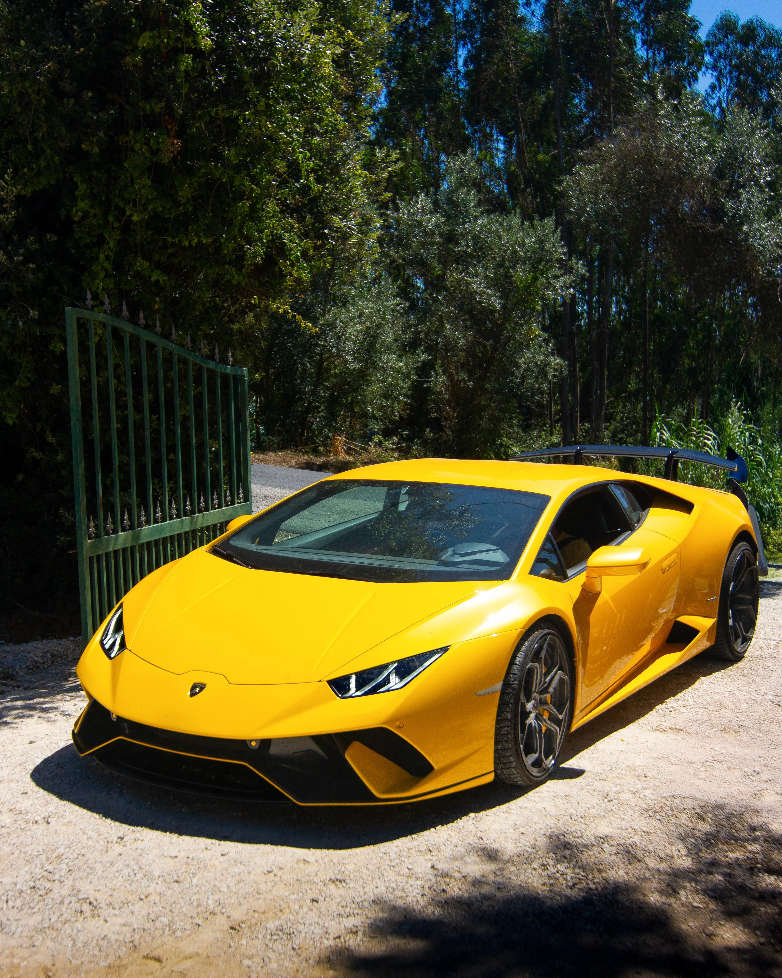 A bright yellow Lamborghini sports car parked near a green gate on a gravel surface, with trees and blue sky in the background.