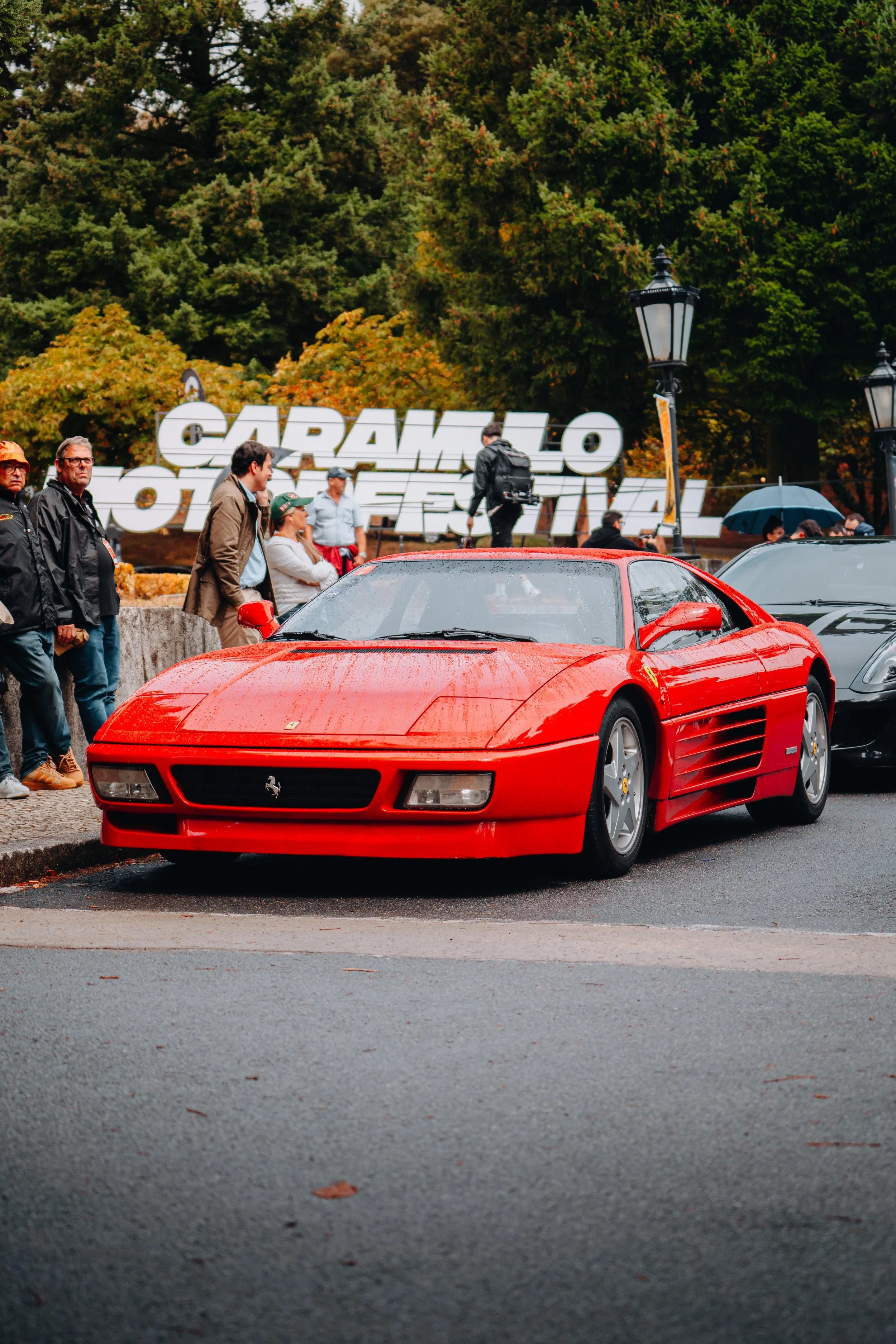 A red Ferrari sports car parked on a street during the Carangelo Motor Festival in fall, with people and large festival sign in the background.