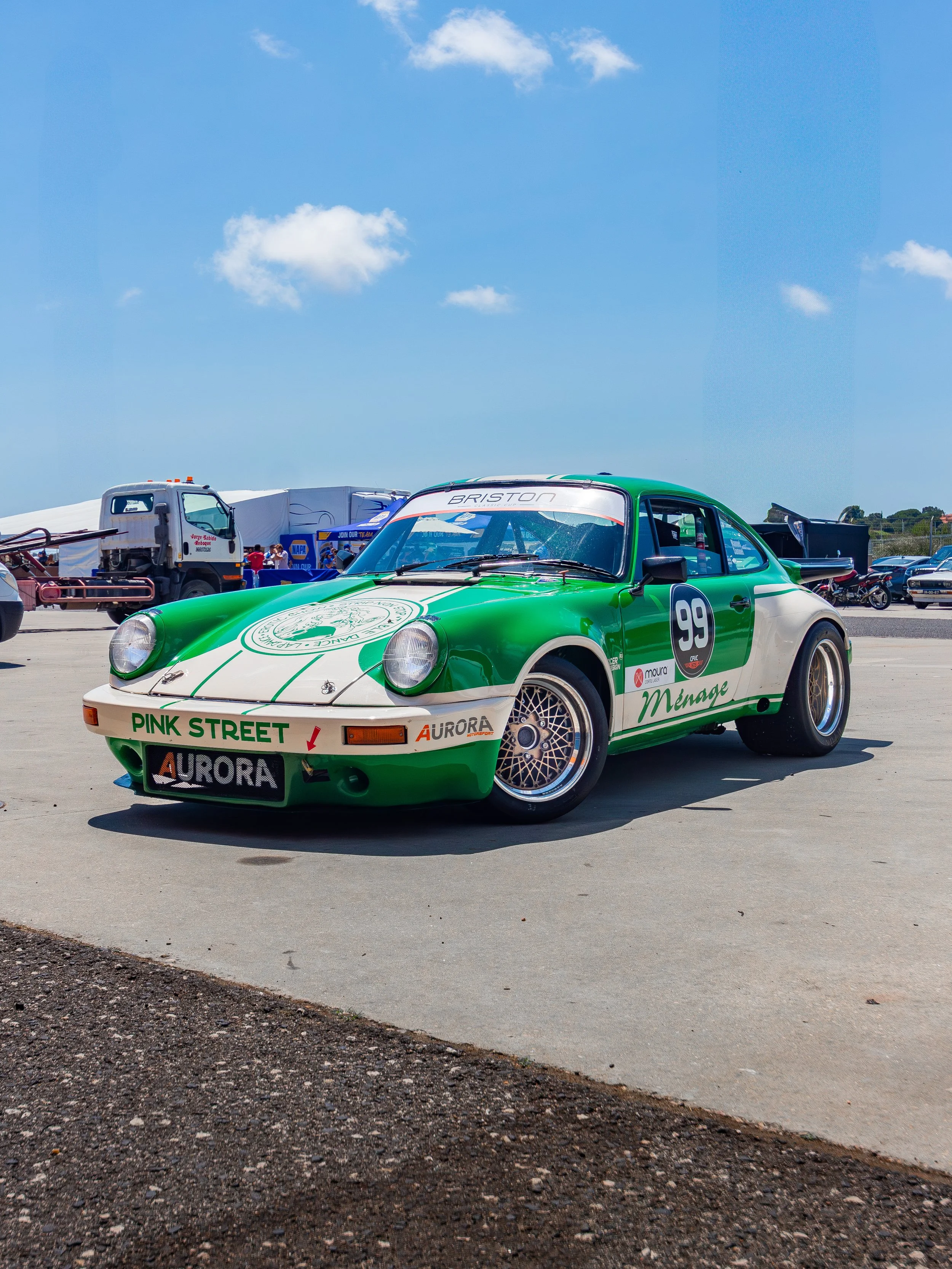 Green and white vintage race car with the number 99 on the side, parked on a tarmac at a car event under a blue sky with a few clouds.