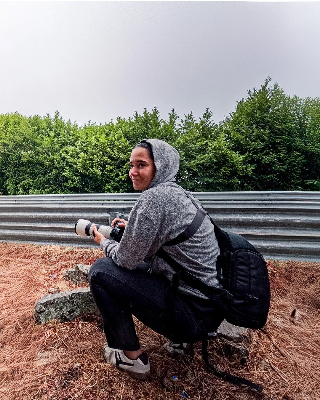 Person crouching outdoors holding a camera with a long lens, wearing a gray hoodie, black pants, and sneakers, with a backpack, beside a metal guardrail and green bushes in the background.