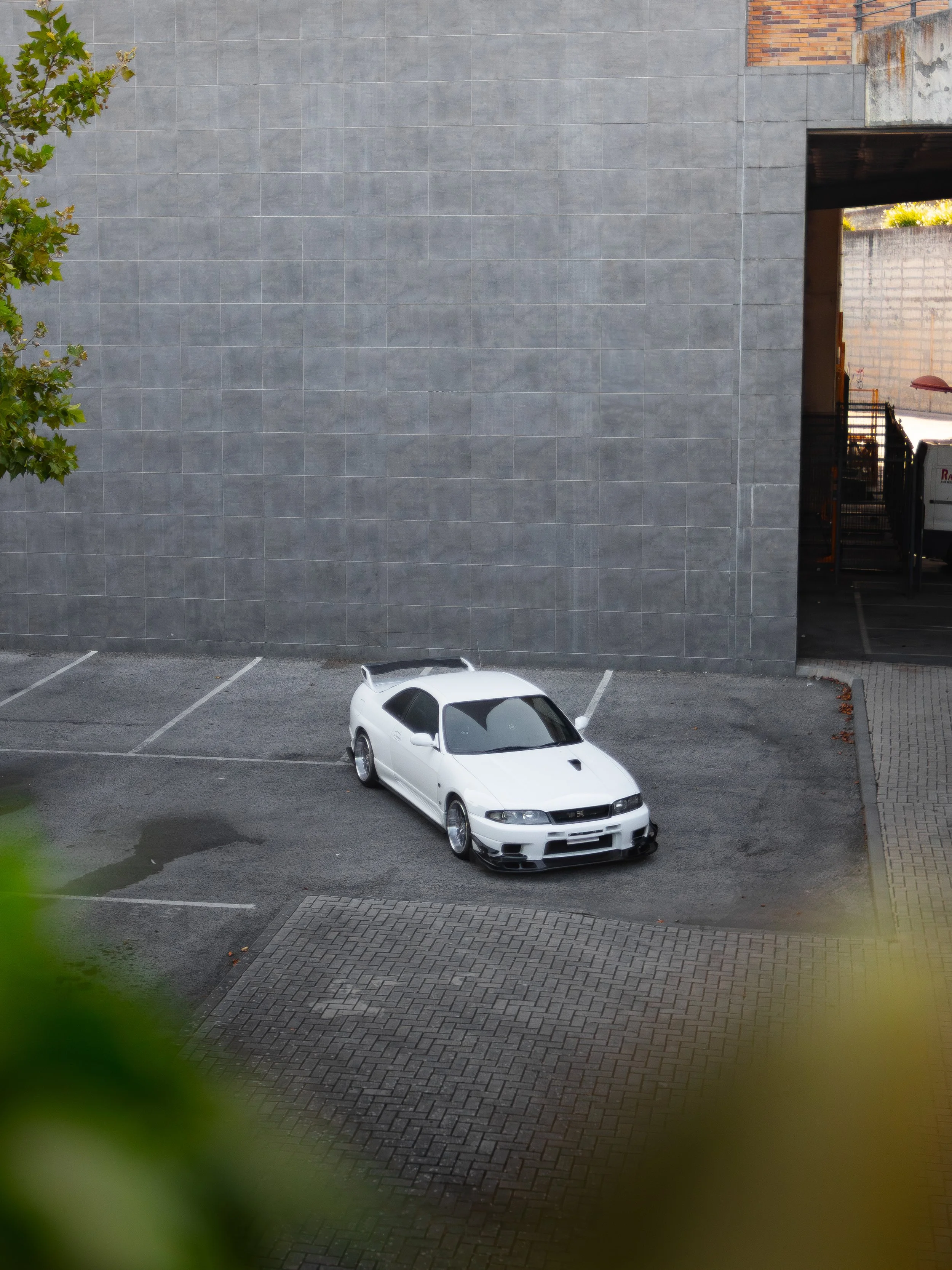 A white sports car parked in an empty parking lot adjacent to a large gray building wall, with a tree partially visible on the left side.
