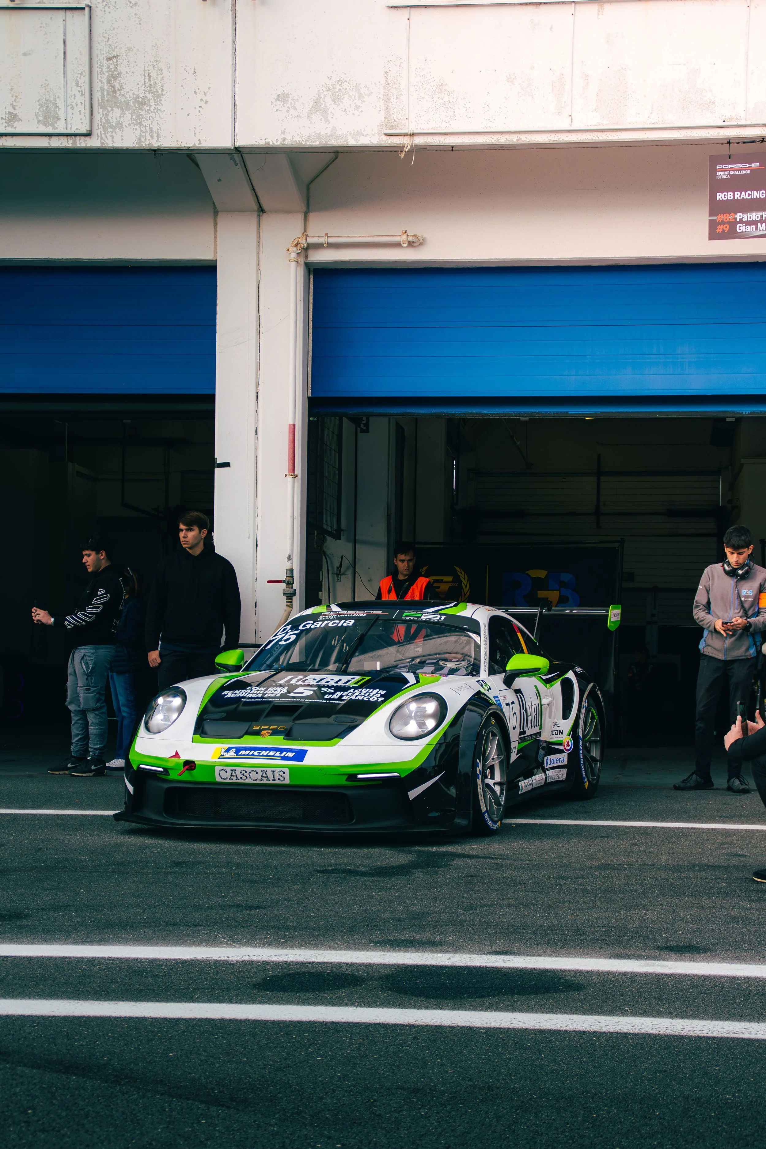 A race car in the pit lane surrounded by people, with a garage door in the background.