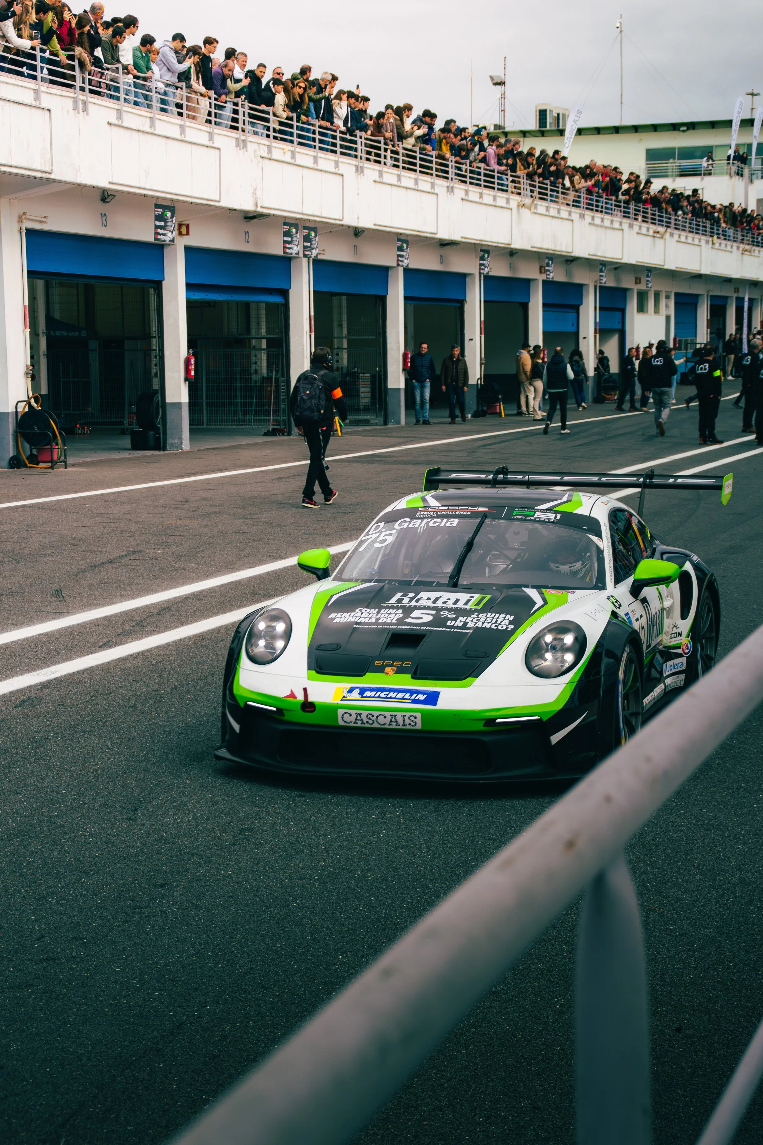 A green and white race car with the number 25 on it, positioned in the pit lane at a race track, with a crowd of spectators watching from the upper stands.