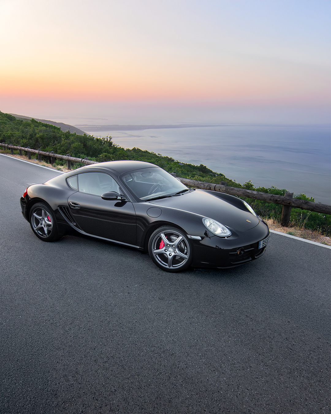A black sports car parked on the side of a coastal road at sunset with the ocean and hills in the background.