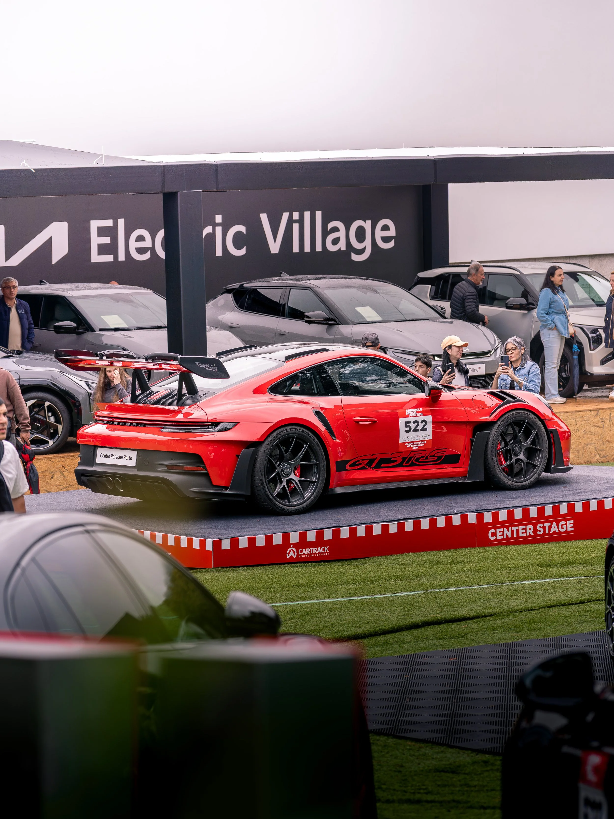 A red Porsche race car on a stage at an auto show, with people around taking photos and observing, behind a sign that reads 'Electric Village'.