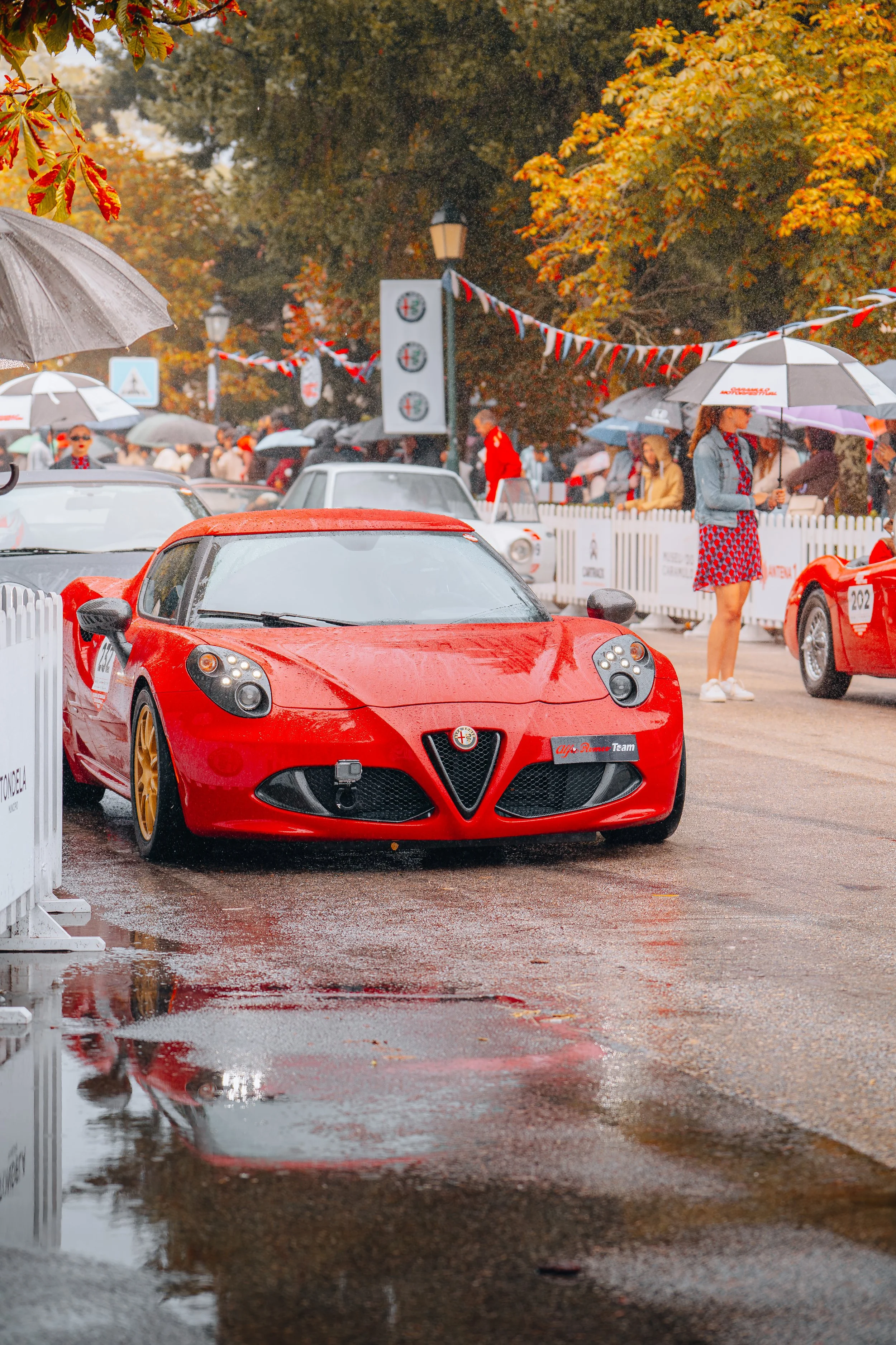 A red sports car parked on a wet street during a rainy day at a car event, with people holding umbrellas and autumn trees in the background.