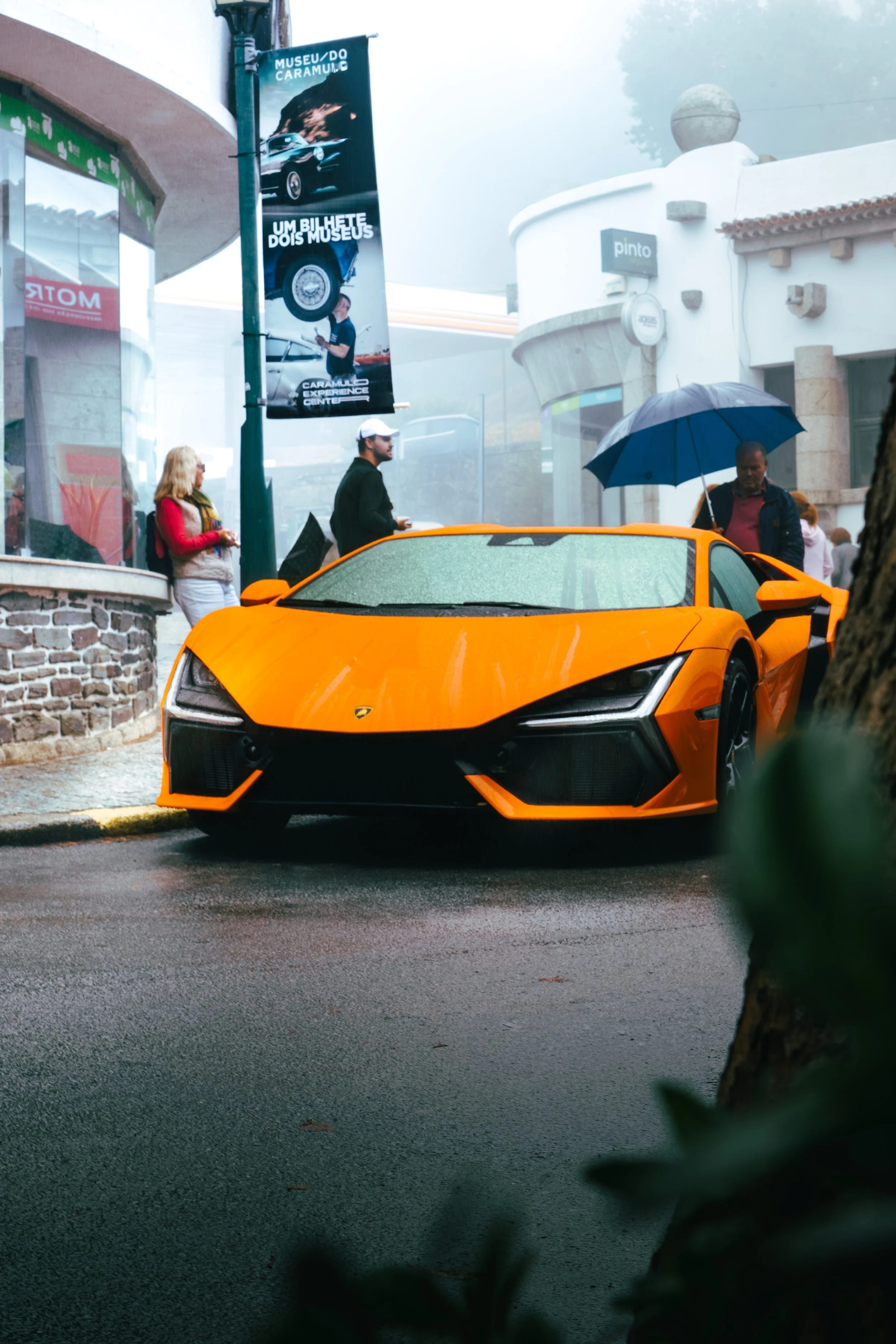 Orange Lamborghini sports car parked on city street on a rainy day, with four people standing nearby, one holding an umbrella, and a building with advertisements in the background.