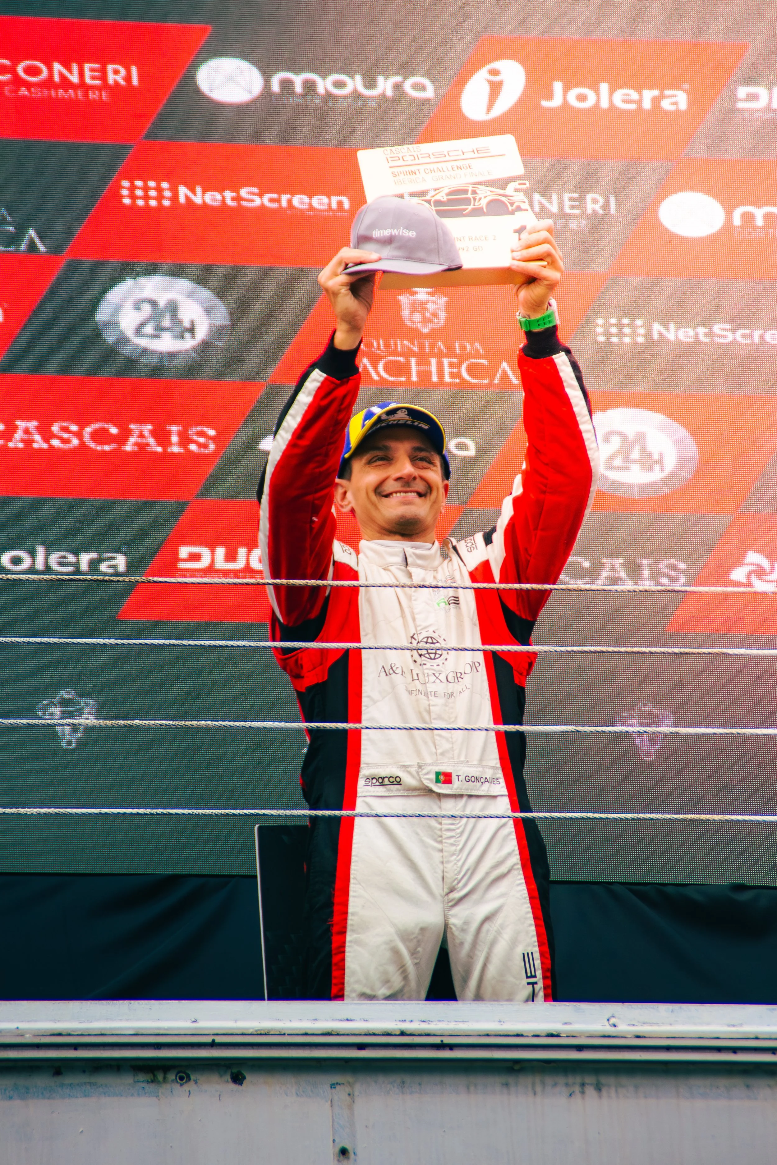 Race car driver standing on the winners' podium, smiling, holding a trophy and a cap above his head, with a checkered background featuring logos of sponsors.