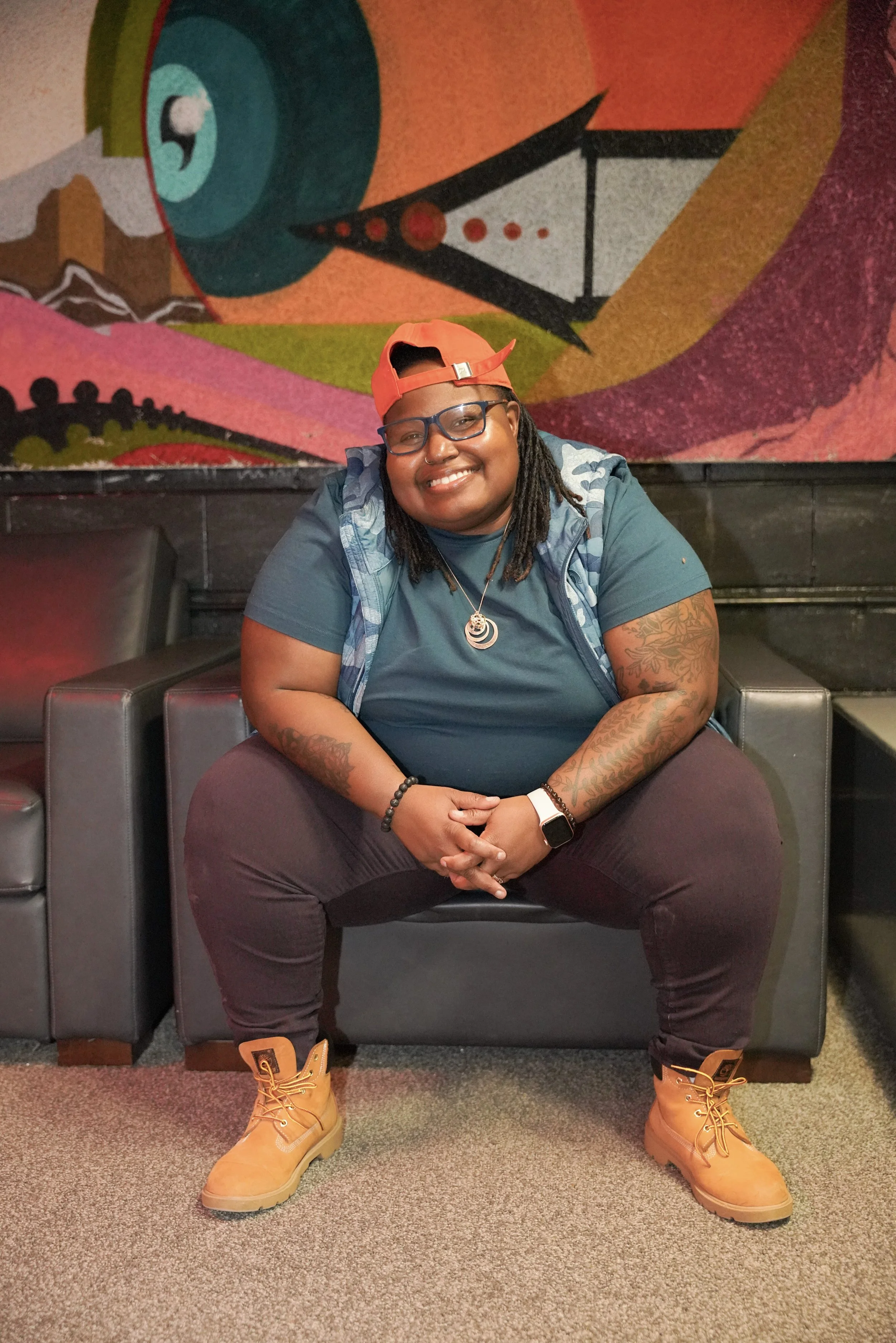 A woman with glasses, dreadlocks, and tattoos, smiling and sitting on a black sofa in front of a colorful abstract mural, wearing a red cap, a teal shirt, yellow work boots, and jewelry.