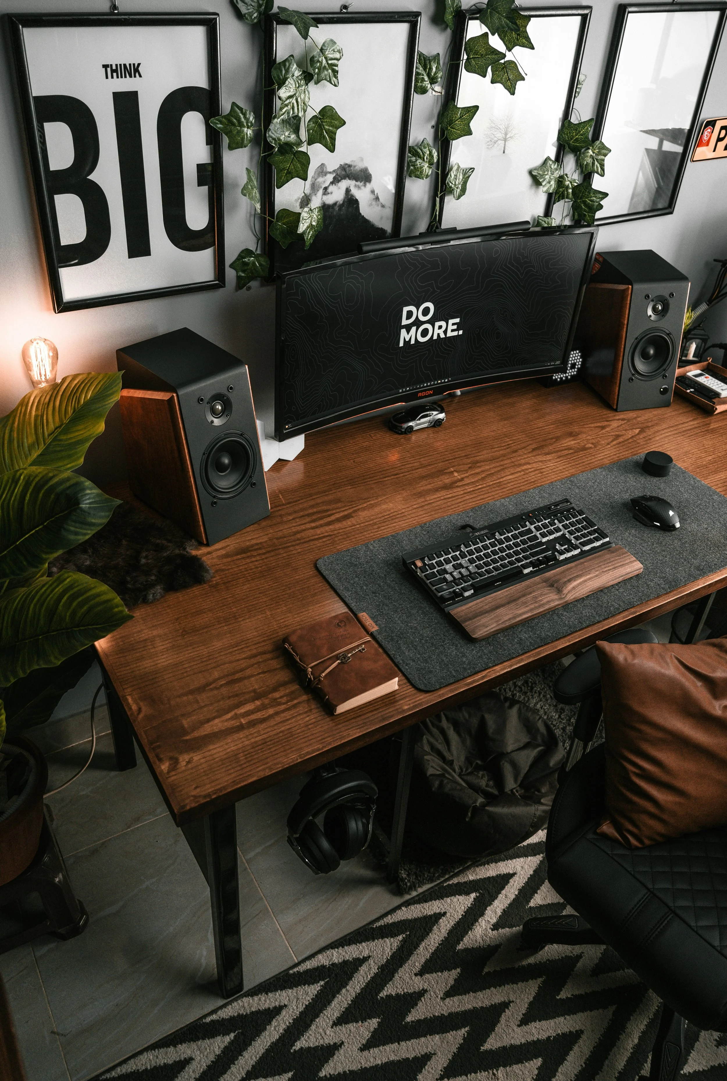 A home office workspace with a wooden desk, two black and brown speakers, a curved monitor, a keyboard, mouse, and a notebook, surrounded by framed prints and green ivy on the wall, a plant, and headphones hanging under the desk.