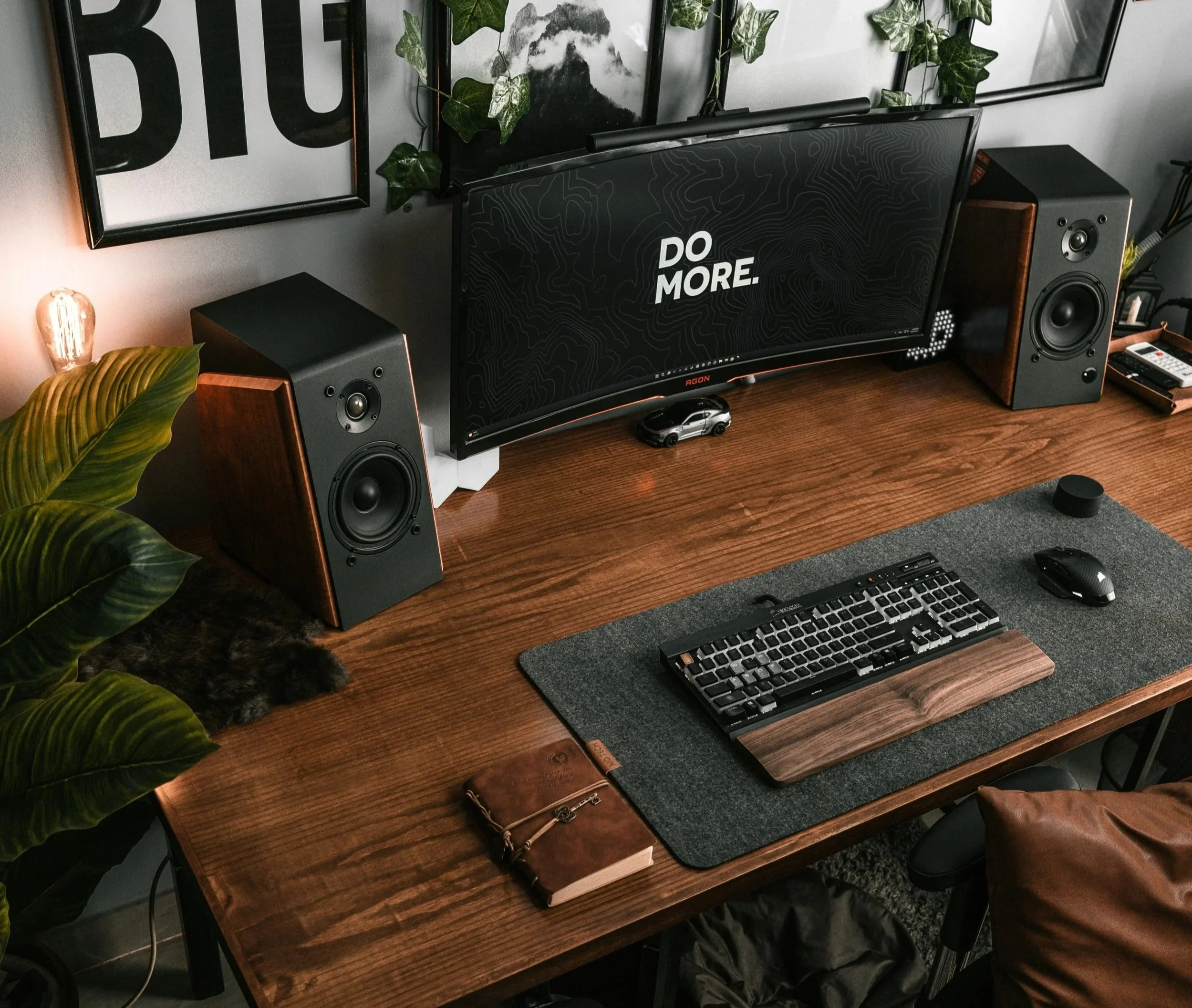 A wooden desk with a computer monitor, a keyboard, mouse, and mouse pad. Two black and wood speakers, a small car model, a plant, framed wall art, a leather wallet, and a book are also on the desk.