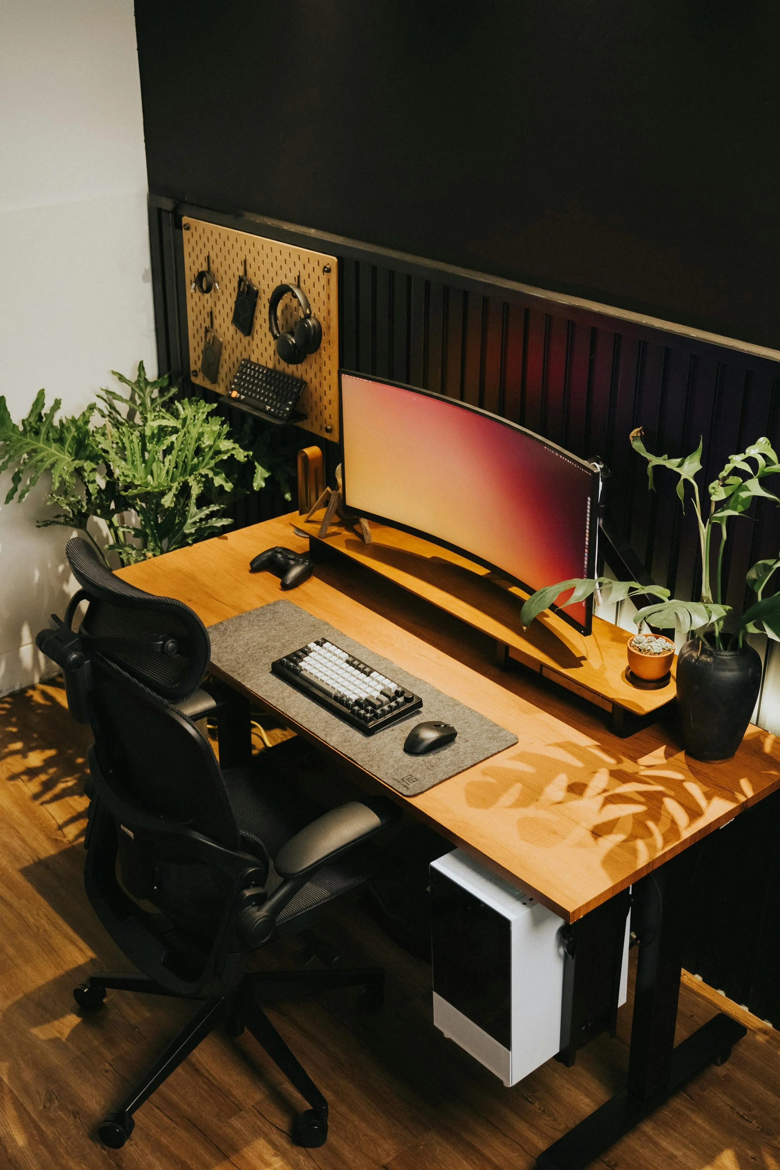 Modern home office with wooden desk, curved computer monitor, keyboard, mouse, ergonomic office chair, and large plants, with a wall-mounted pegboard holding headphones, a keyboard, and hooks.