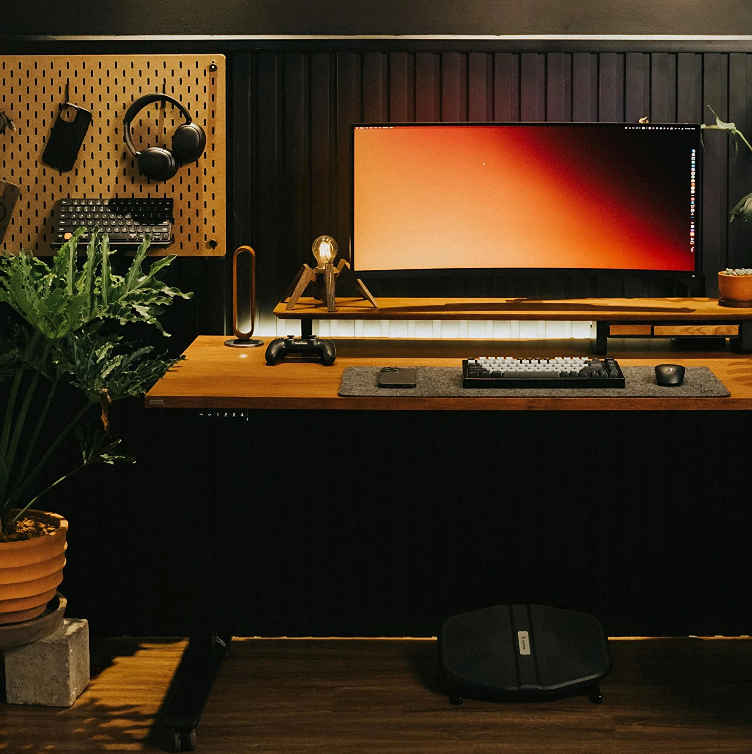 A modern desk with a large monitor, keyboard, mouse, and a game controller, surrounded by plants and decorative objects, with a pegboard holding headphones, a phone, and a small keyboard in the background.