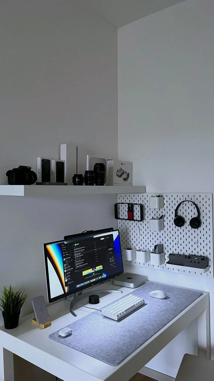 A modern workspace with a white desk, a desktop computer, a small potted plant, and various tech accessories on a pegboard behind the desk. The shelf above holds cameras and camera lenses.