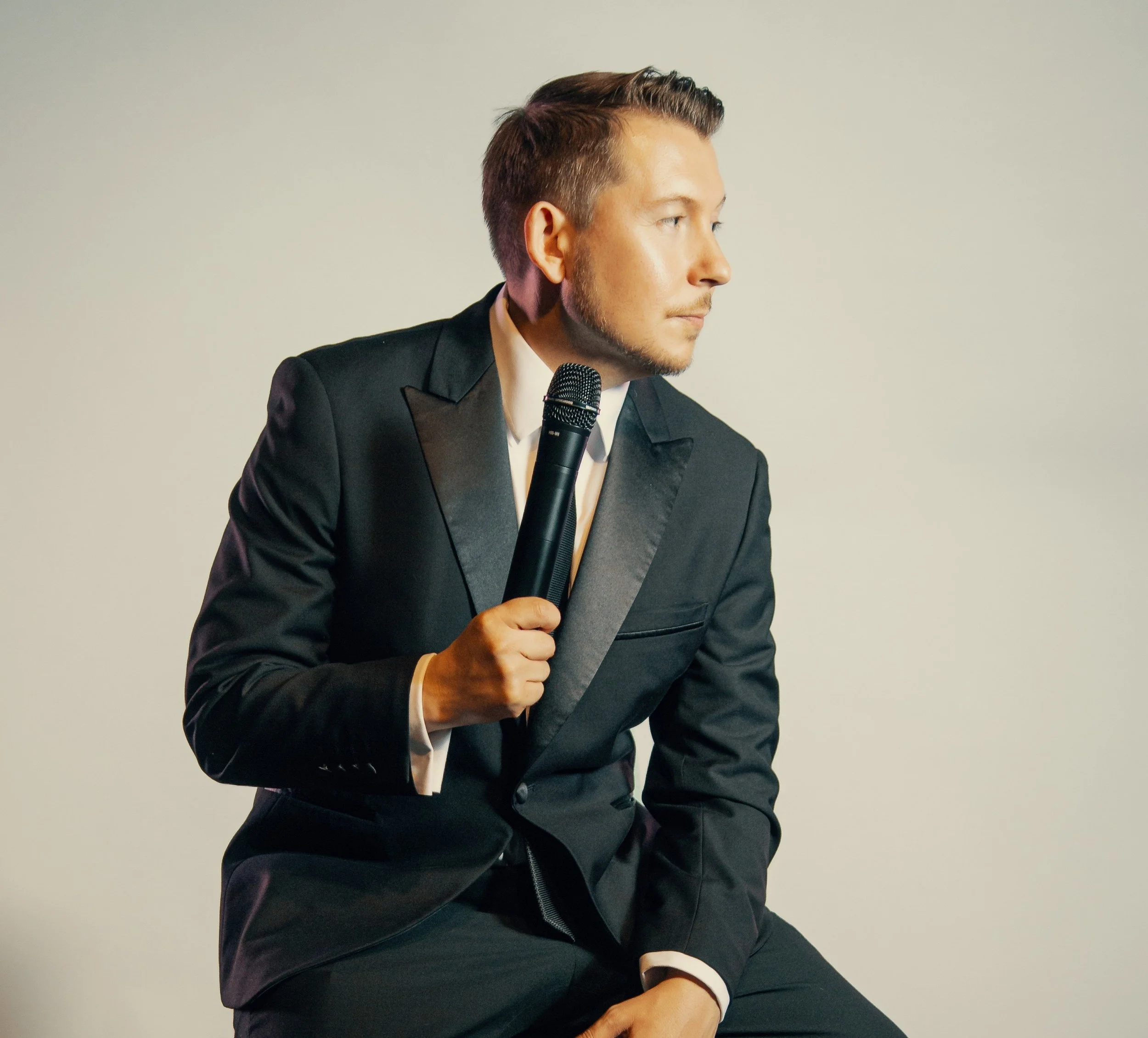 A man in a black tuxedo holds a microphone and looks to the right, sitting against a plain light-colored background.