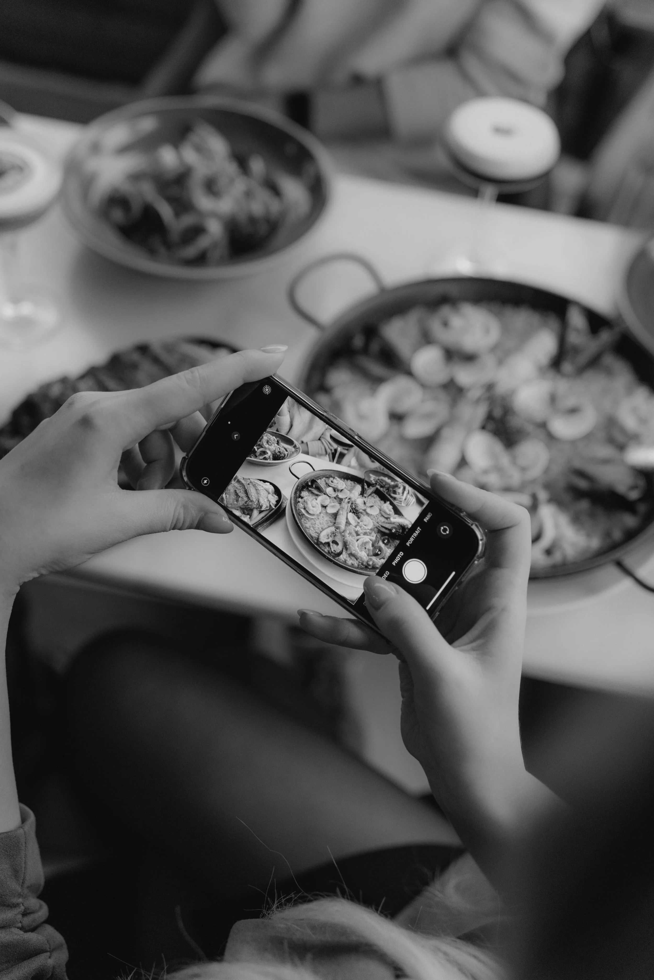 A person is taking a photo of a table filled with various dishes of food with a smartphone in a black-and-white photo.