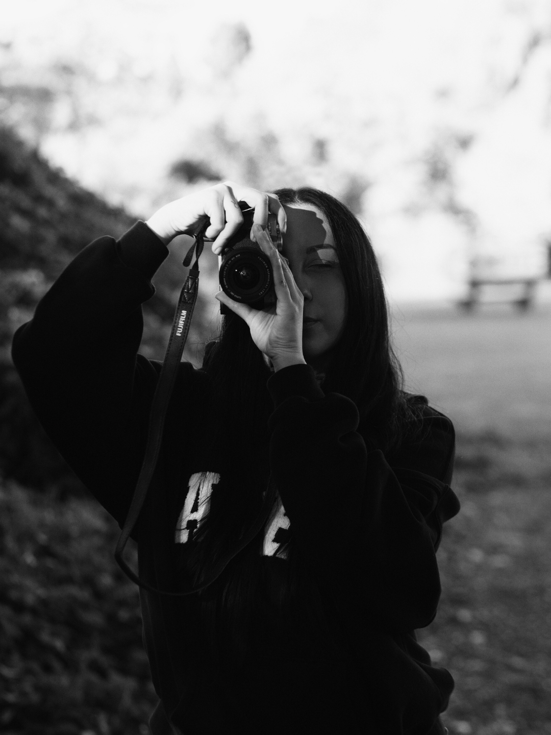 A woman with long dark hair holding a camera, taking a photo outdoors, in a black and white image.