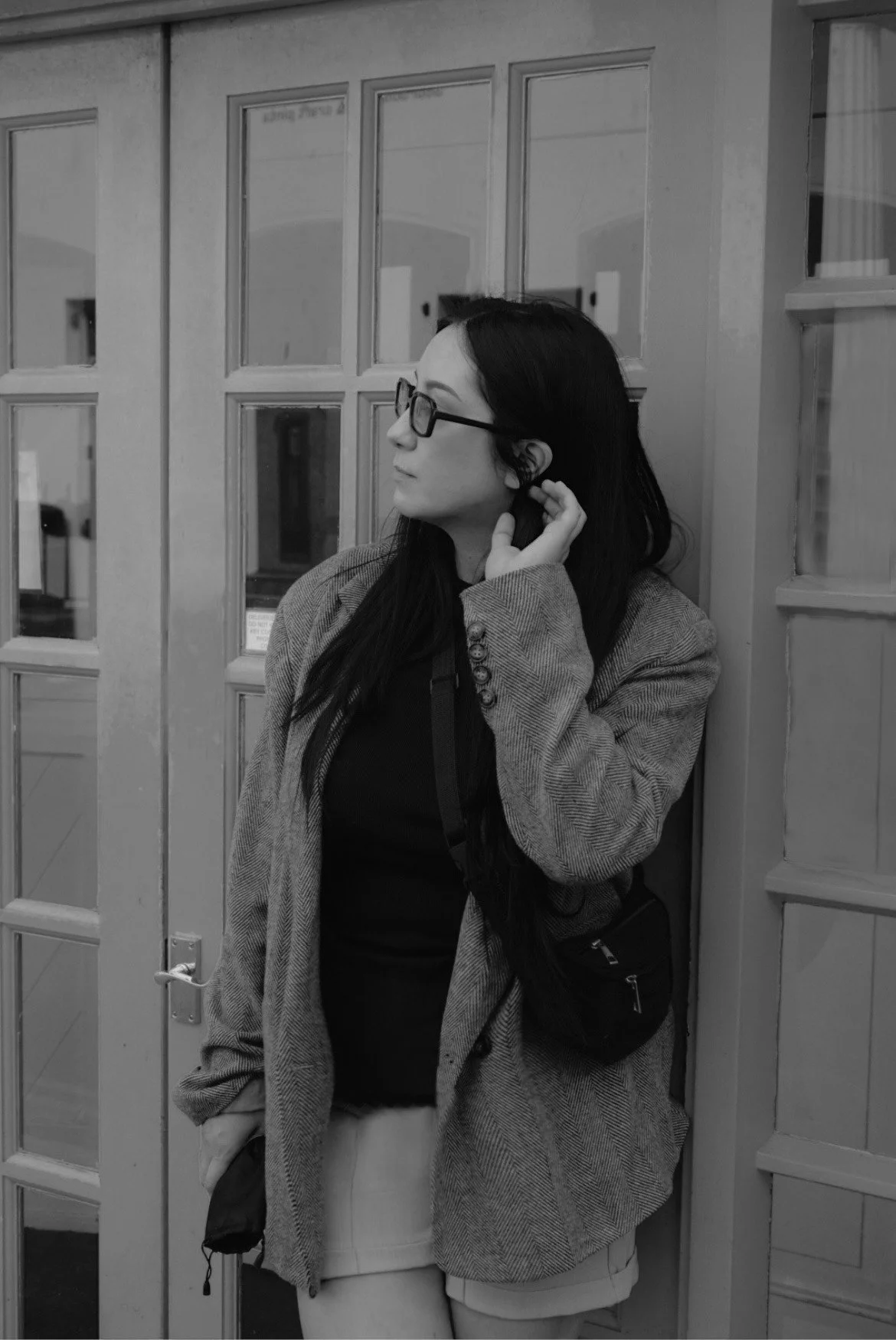 A woman wearing glasses, a patterned blazer, and light-colored shorts, standing against a wooden door, adjusting her earring.