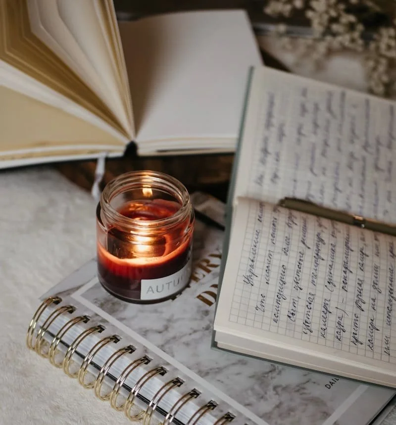 An open notebook with handwritten notes, a closed book, a pen, a lit candle in a glass jar, and a spiral-bound notebook on a marble surface.