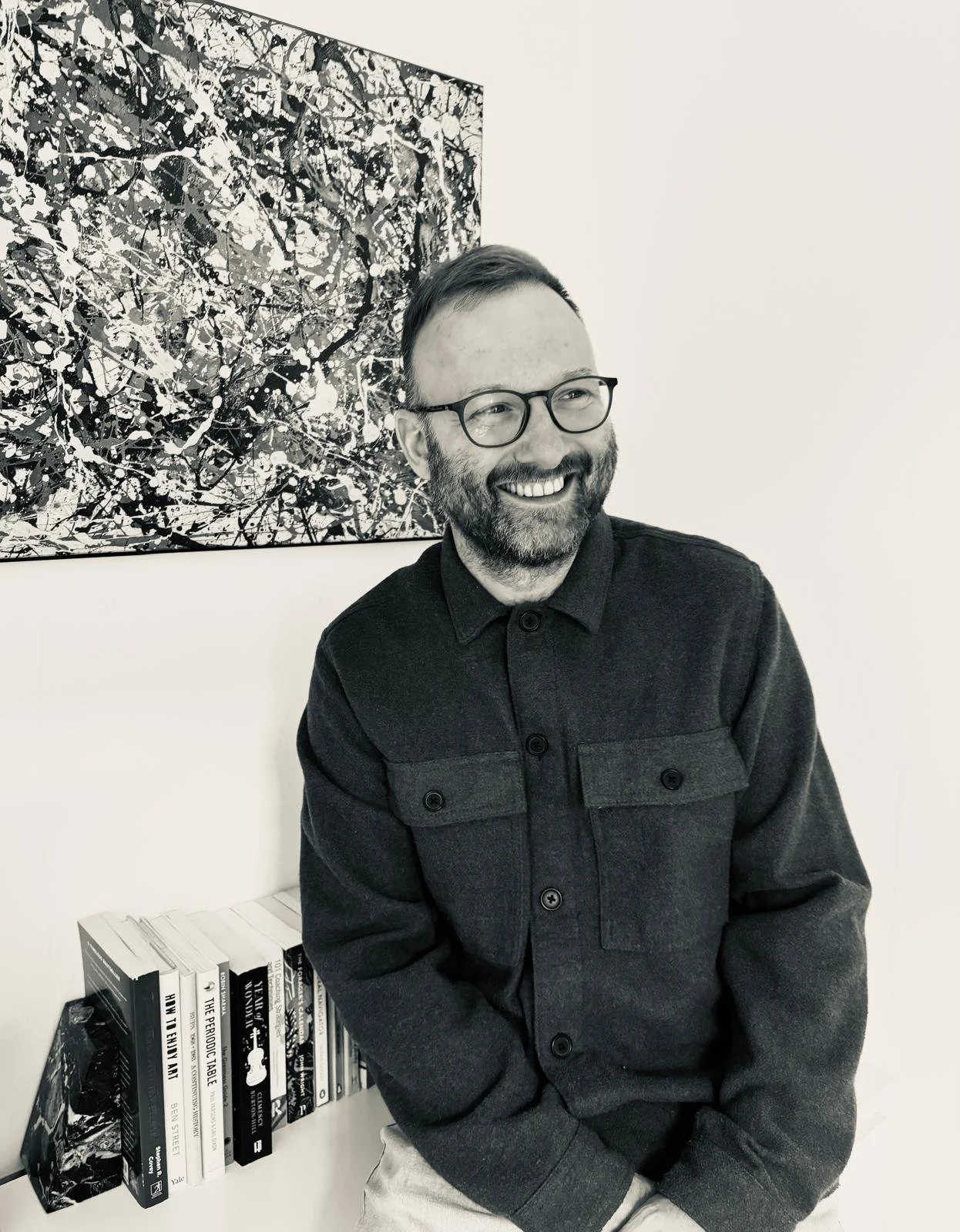 A man with glasses and a beard smiling, sitting in front of a bookshelf with books, and a large abstract black and white painting on the wall behind him.