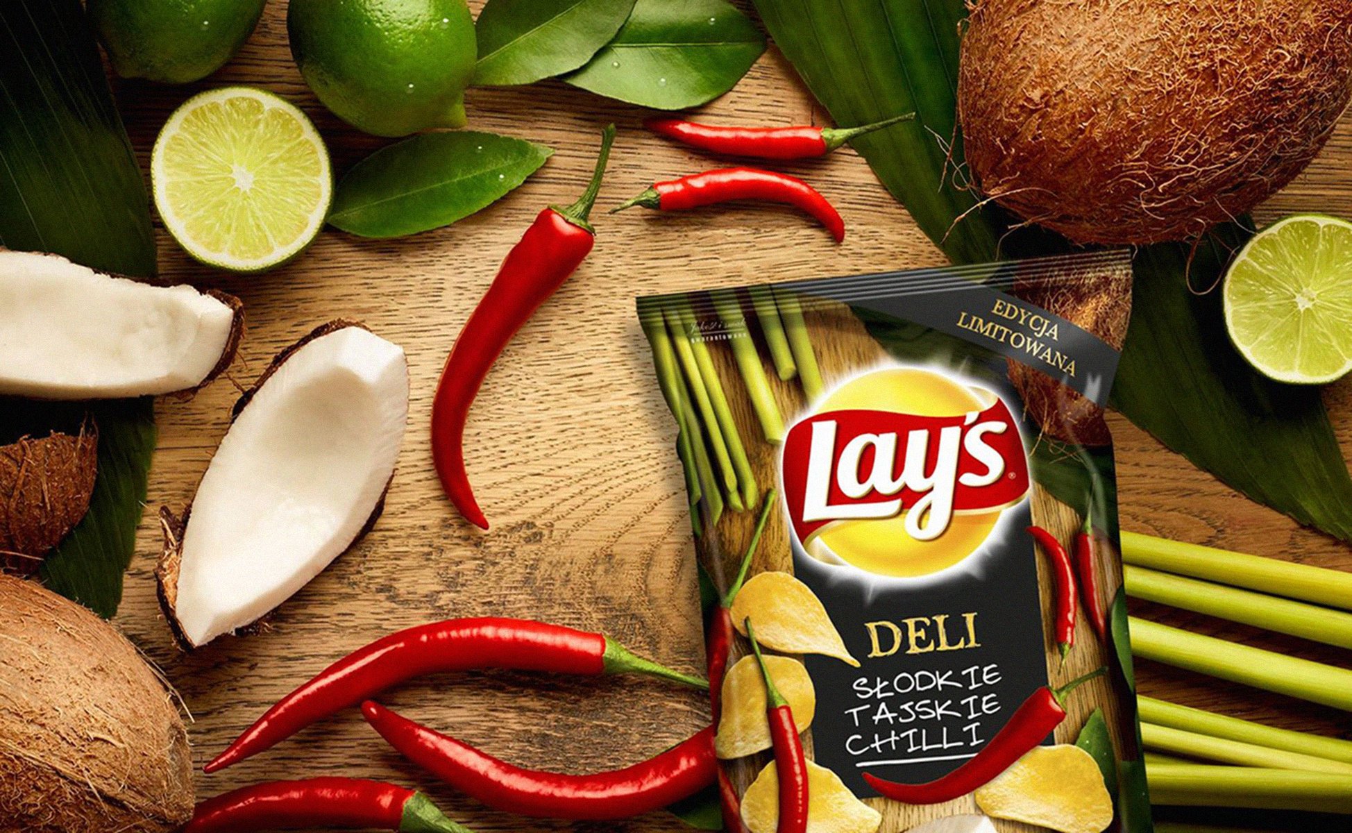 A packet of Lay's Deli snack chips surrounded by coconut, lime, red chili peppers, coconut shells, green leaves, and stalks of lemongrass on a wooden surface.