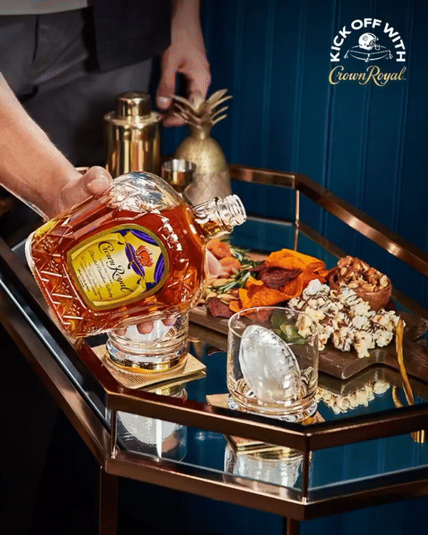 Person pouring Crown Royal whiskey into a glass on a bar cart with snacks and decor in the background.