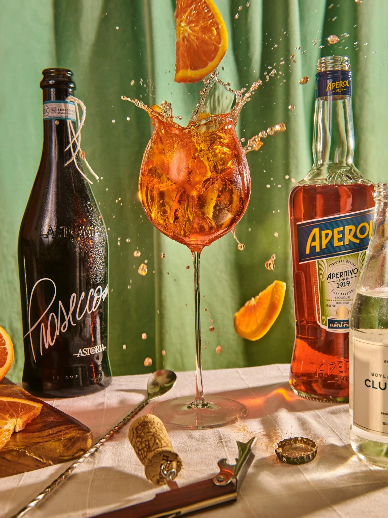 A cocktail glass with an orange slice in it, splashing as a liquid is added, surrounded by liquor bottles, an orange slice on the table, and a corkscrew on a white tablecloth with a green curtain background.