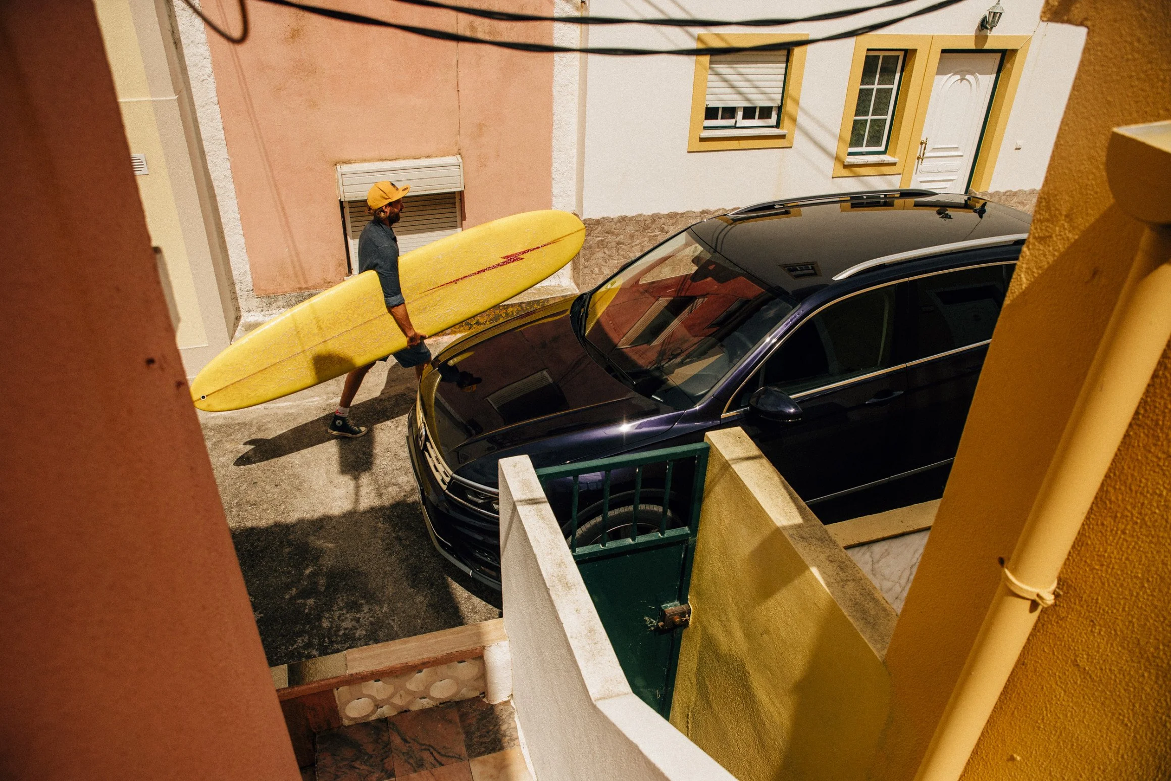 Man carrying a yellow surfboard walking past a black car parked in a courtyard between buildings with pink, yellow, and white walls.