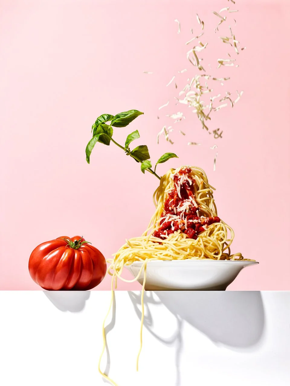 Spaghetti pasta with tomato sauce, basil leaves, grated cheese, next to a large heirloom tomato, with cheese being sprinkled onto the pasta.