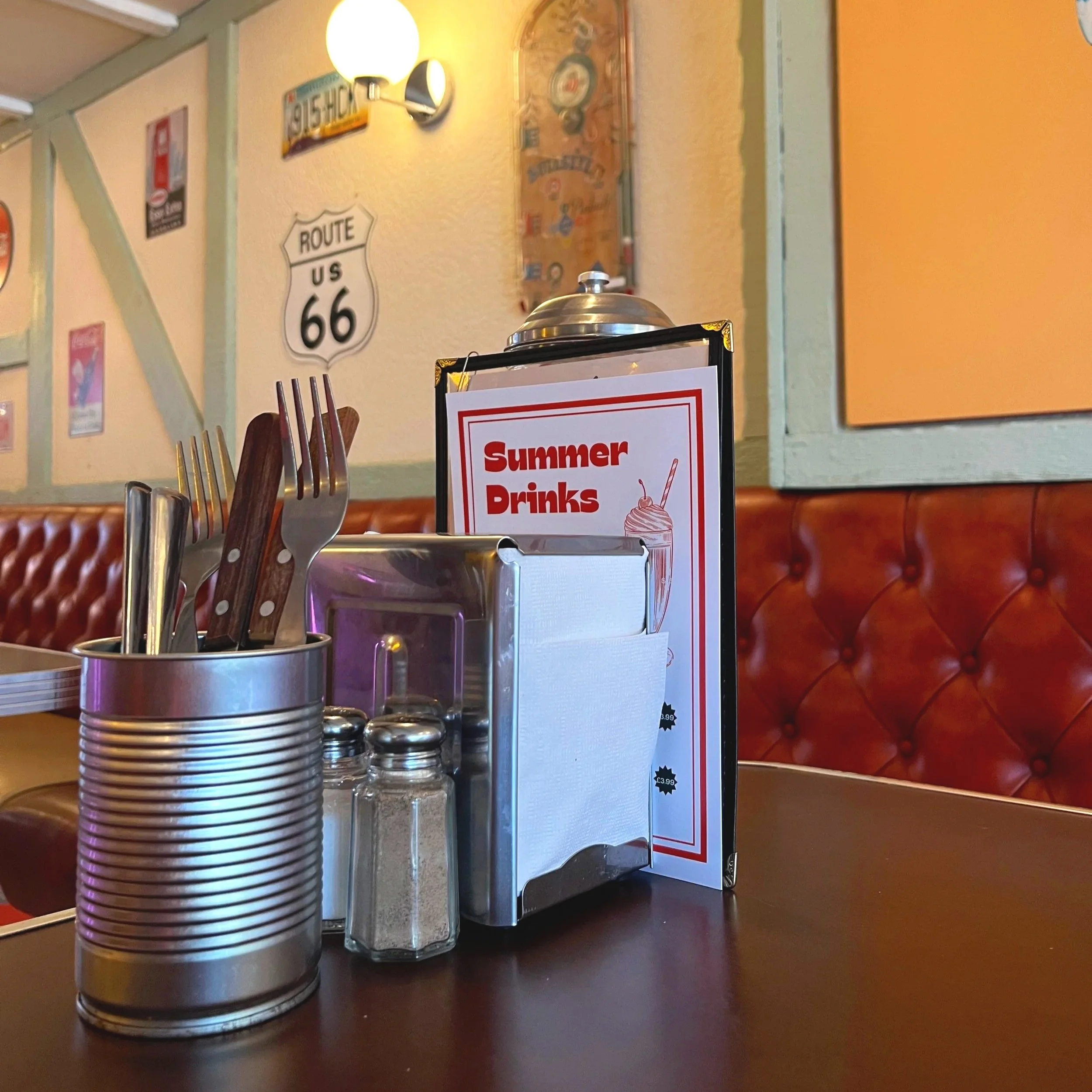 Table setting at a restaurant with utensils, salt and pepper shakers, a condiment holder, and a summer drinks menu, with a booth and Route 66 sign visible in the background.