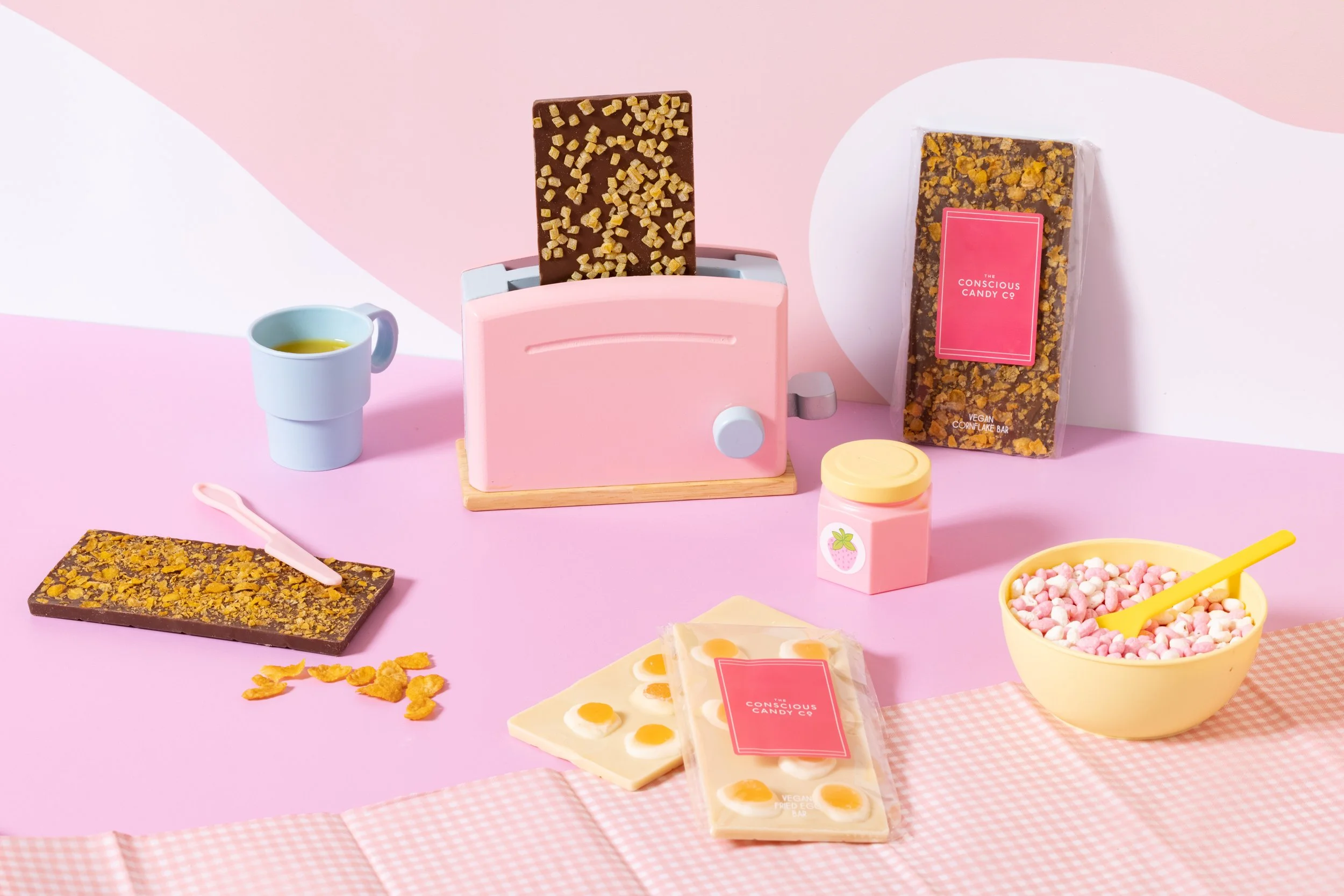 A pink kitchen scene with a toy toaster, chocolate bars, a bowl of cereal, a cup of tea or coffee, a pink jar, and packages of vegan candy, set against a pink and white background.