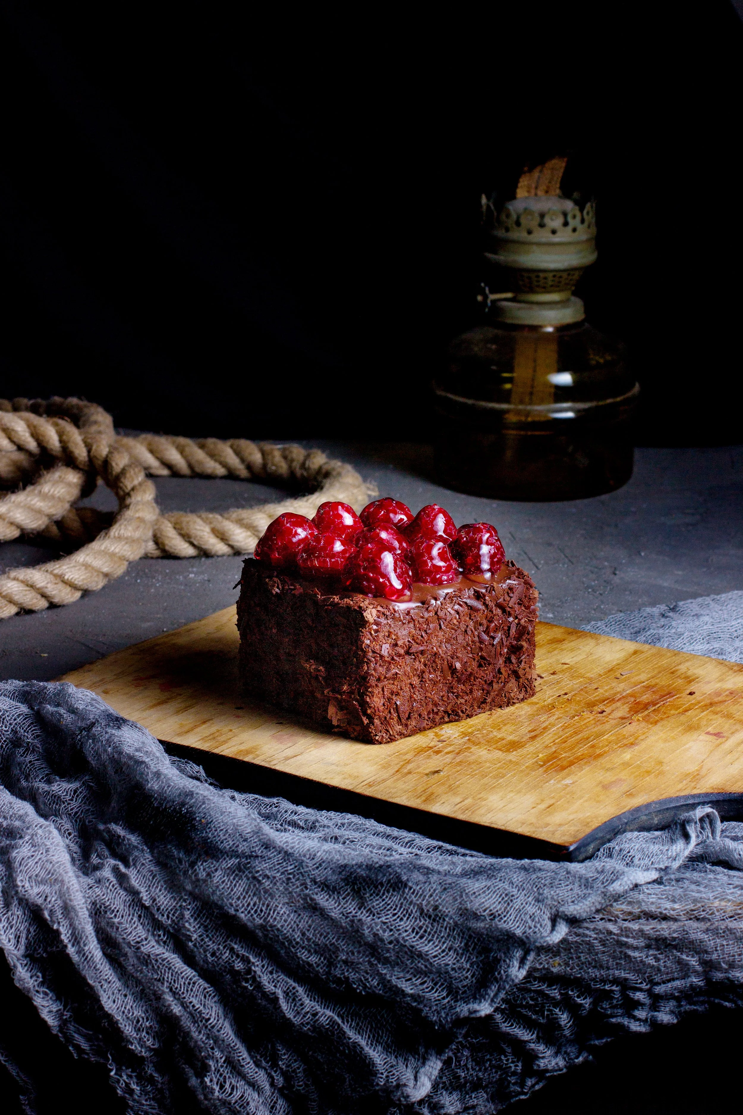 A piece of chocolate cake topped with red berries on a wooden cutting board, with a blue cloth underneath and a dark background with an old oil lamp and a thick rope.