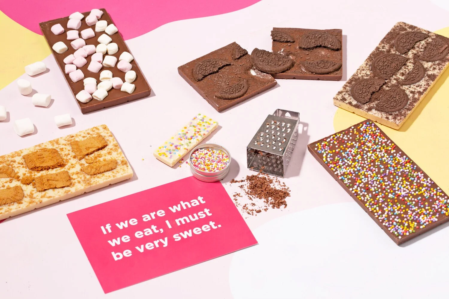 Various types of chocolate bars, cookies, sprinkles, and toppings on a pink and yellow background. A pink card reads, 'If we are what we eat, I must be very sweet.'