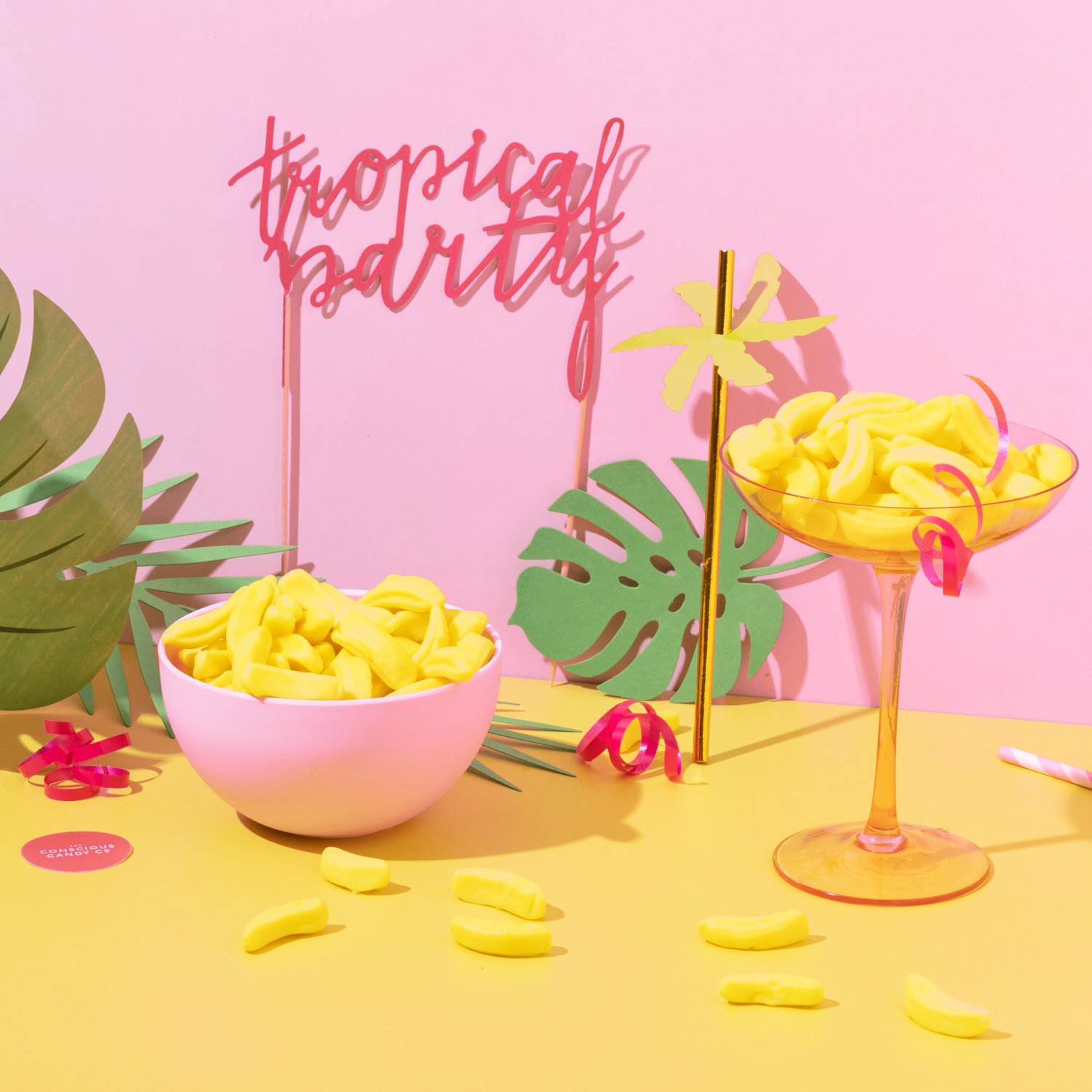 Decorative tropical-themed setup with pink aloha-style sign reading 'tropical party,' yellow gummy candies in a white bowl and a yellow cocktail glass, green paper palm leaves, and pink and yellow party decorations on a pink and yellow background.
