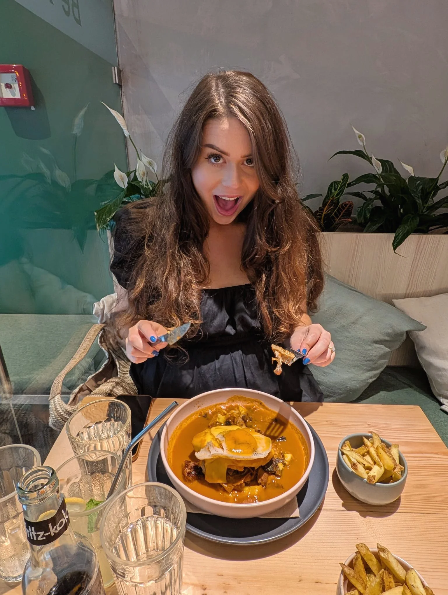 A young woman with long brown hair, wearing a black dress, sitting at a table in a restaurant. She is excitedly holding a piece of food and smiling with her mouth open. In front of her is a bowl of food with fried eggs on top, and to the side are bowls of fries.