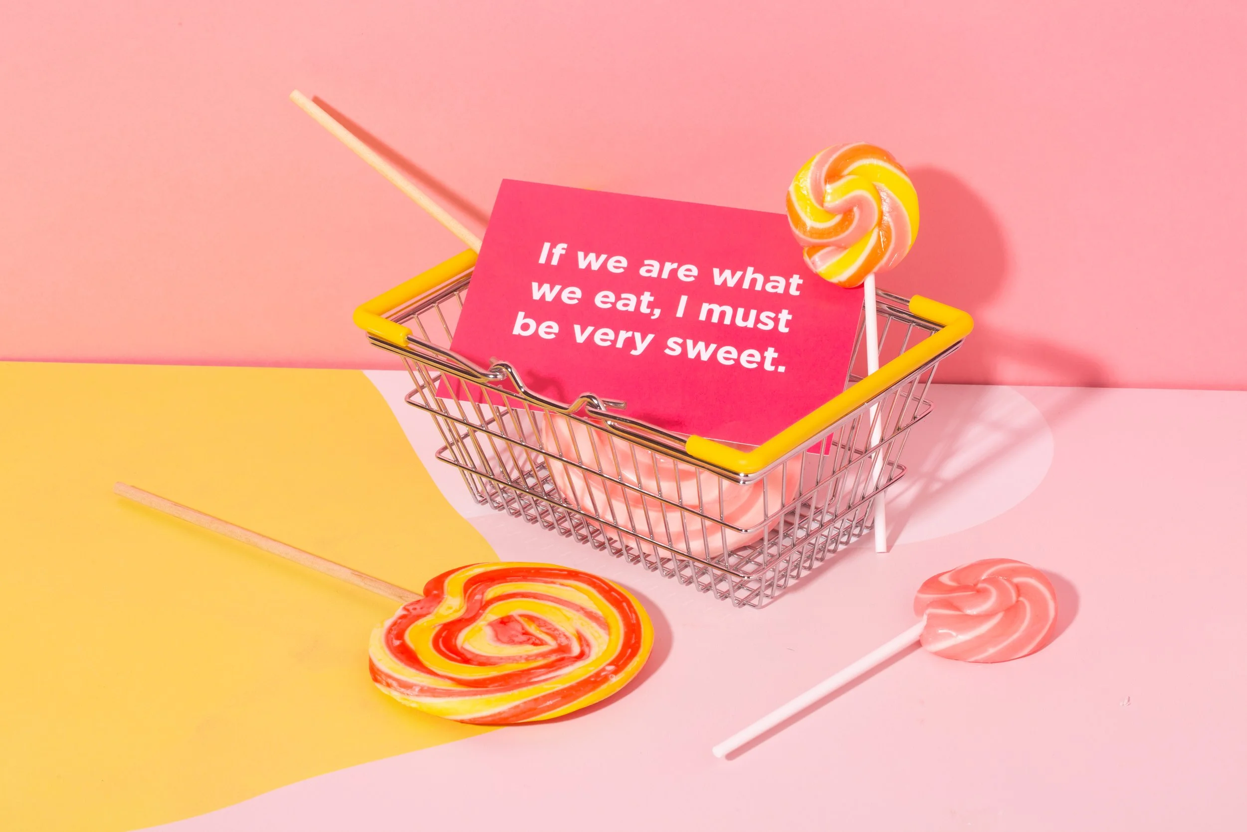 Colorful swirl lollipops in a small shopping cart with a pink sign that reads, 'If we are what we eat, I must be very sweet,' against pink and yellow background.