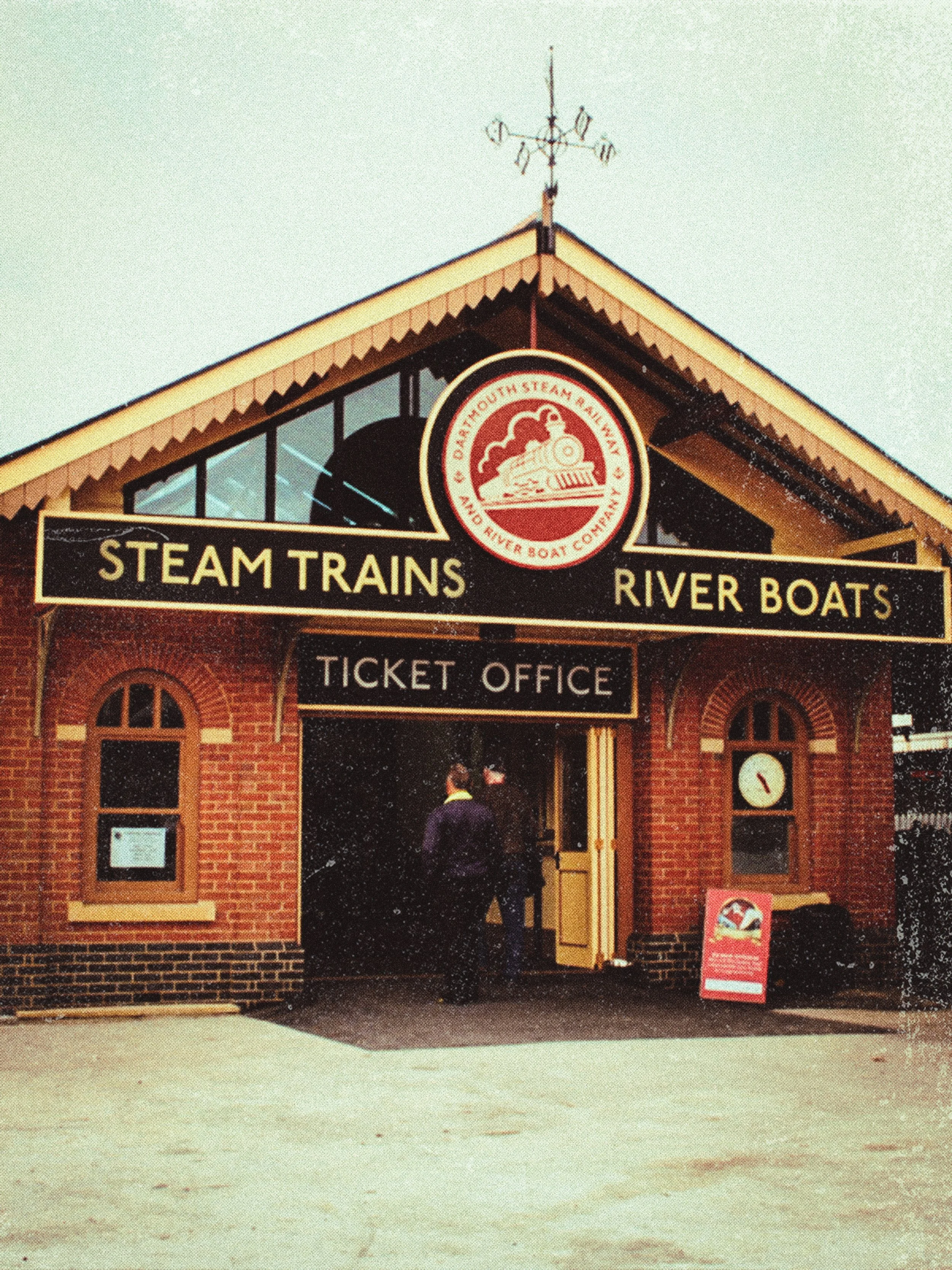 A brick ticket office building for steam trains and river boats with a sign and clock, and people entering.