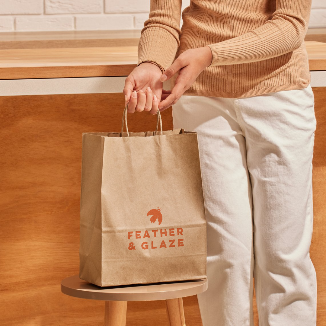 Person holding a brown paper bag with 'FEATHER & GLAZE' logo on it, standing next to a small round table. The person is wearing a tan sweater and white pants, in a setting with a wooden wall and counter.