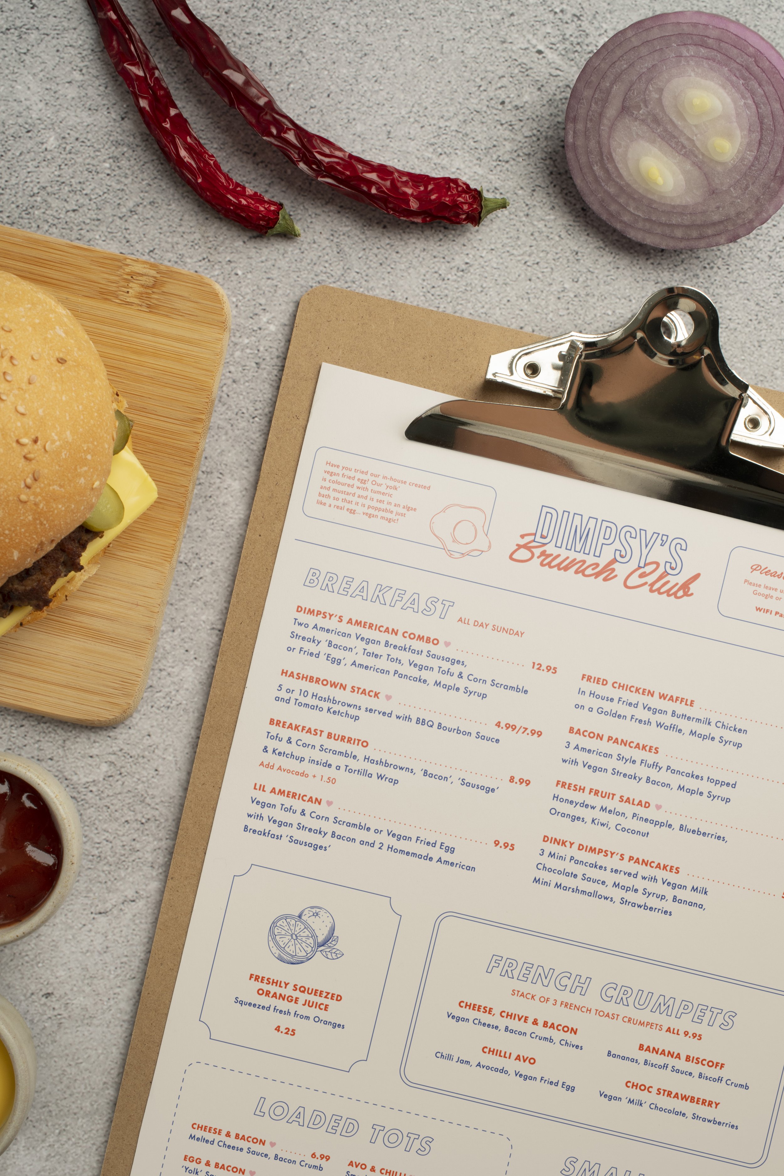 A menu on a clipboard, a burger on a wooden plate, sliced red onion, red chili peppers, and a small bowl of ketchup on a gray table.