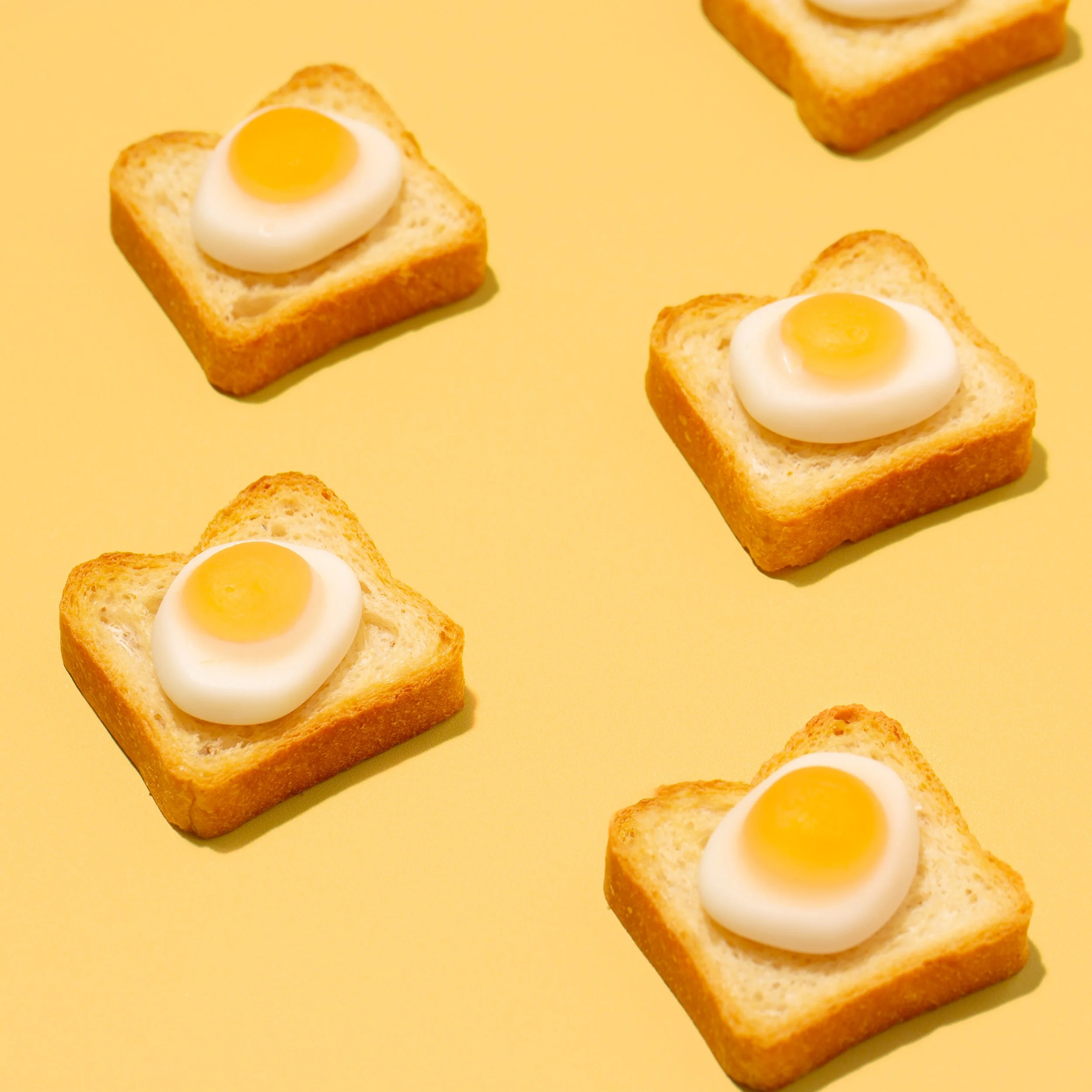 Four slices of toasted bread each topped with a dollop of mayonnaise and a small yellow egg yolk, arranged on a yellow background.
