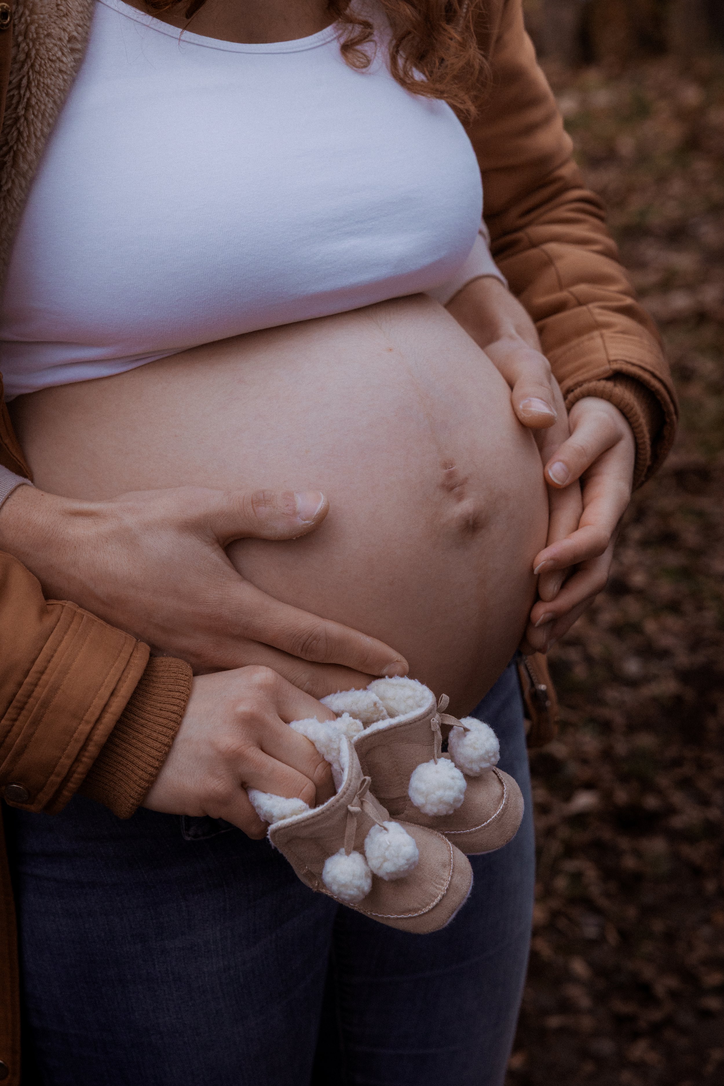 Grossesse d'une femme enceinte tenant ses petits bottillons beige avec pompons blancs devant son ventre nu, en extérieur.