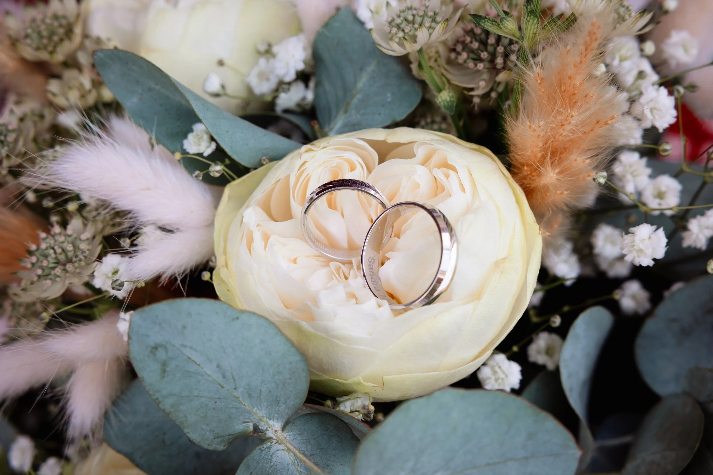 Deux alliances en argent reposant sur une rose blanche entourée d'œillets, de fougères sèches, de plumes et d'autres fleurs blanches.
